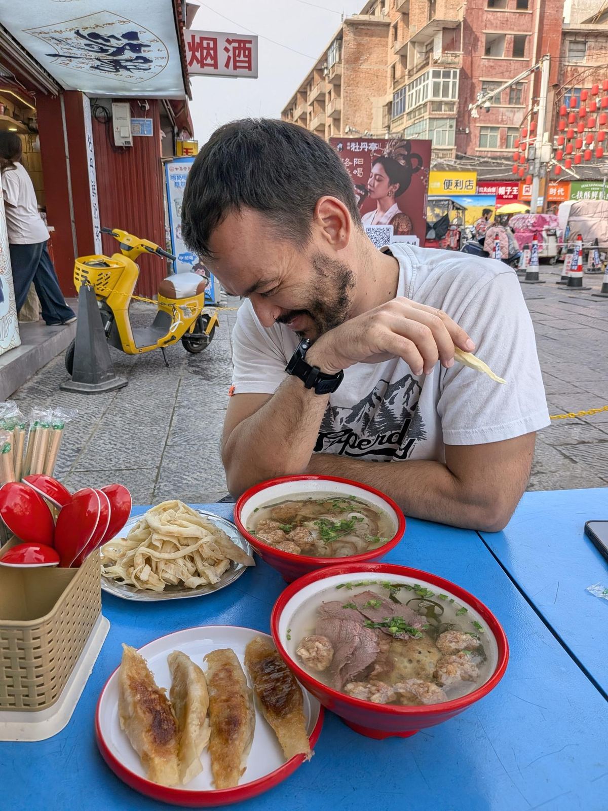 Tristan devant ses deux bols de niurou tang avec leurs brochettes, raviolis frits et tofu à la table extérieure, lanternes rouges au-dessus