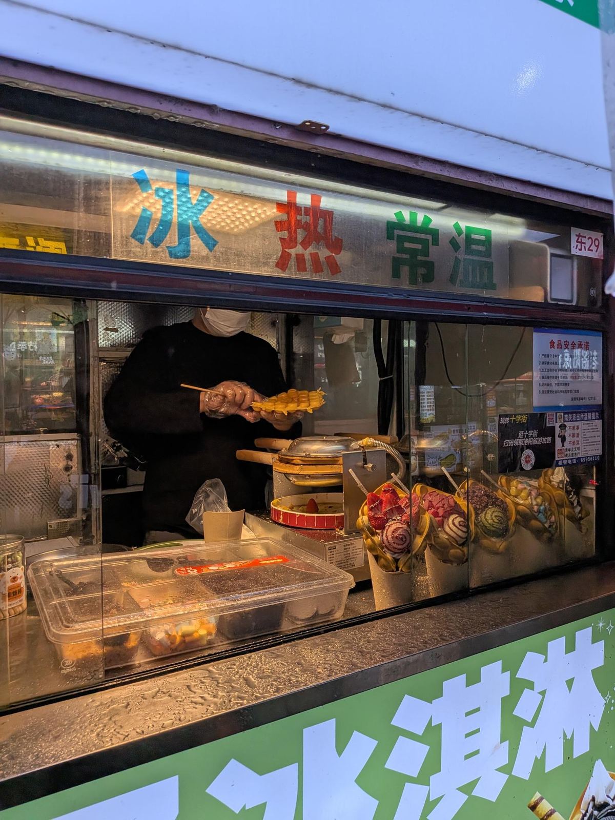 Le stand de gaufres, vendeur en masque qui lève sa gaufre dorée au-dessus du fer, vitre 冰/热/常温, pancarte 食品安全 et QR de la police locale