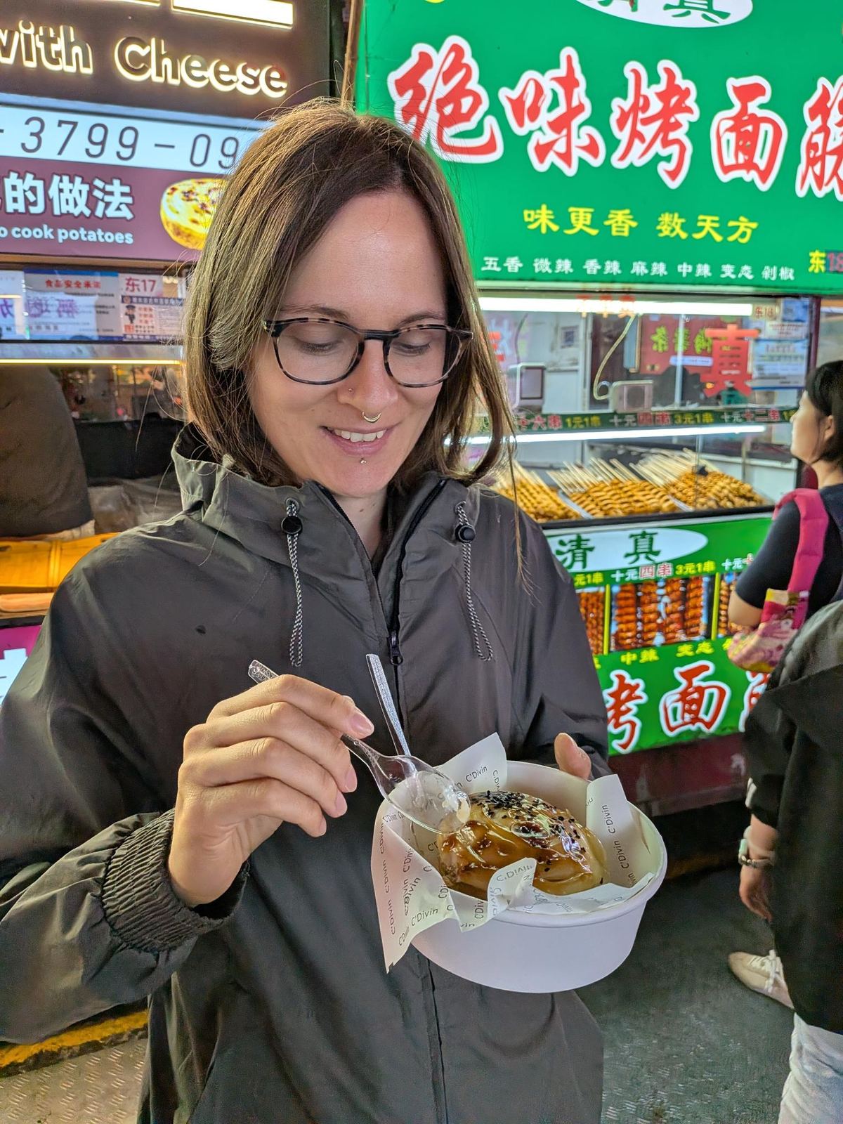 Marion avec son bol de dessert au caramel et aux graines, cuillère levée, stand 绝味烤面筋 halal et ses niveaux de piment en arrière-plan