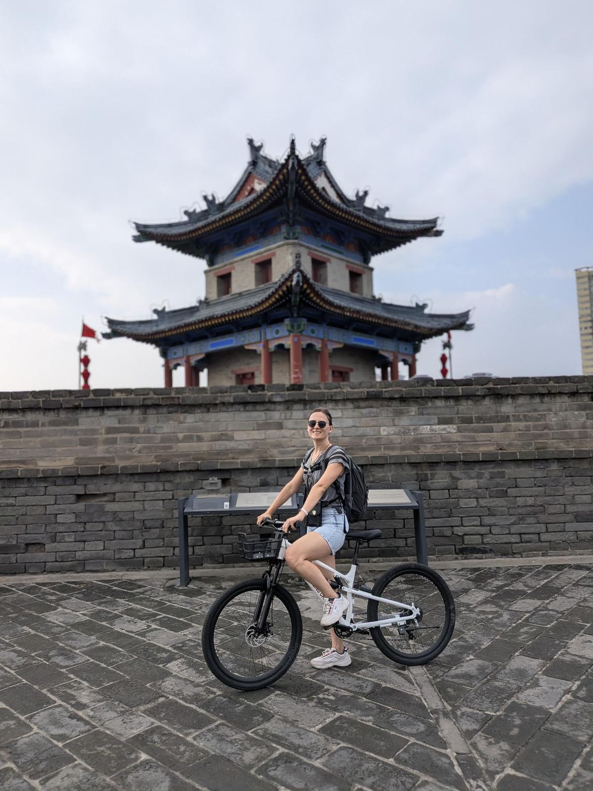 Marion souriante en shorts denim sur son vélo blanc devant le rempart gris, pavillon rouge à toit traditionnel derrière, drapeau rouge à gauche