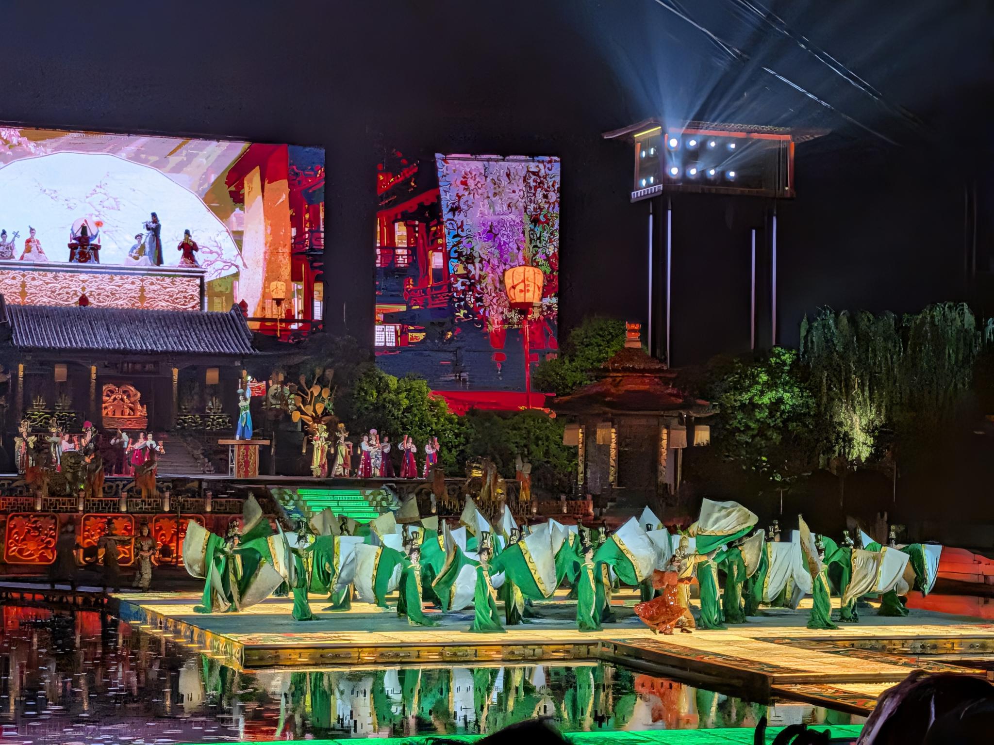 Danseurs en costumes verts et blancs soulevant de grands panneaux de tissu blanc, pavillon à toit tuilé et écrans LED de l'intérieur du palais