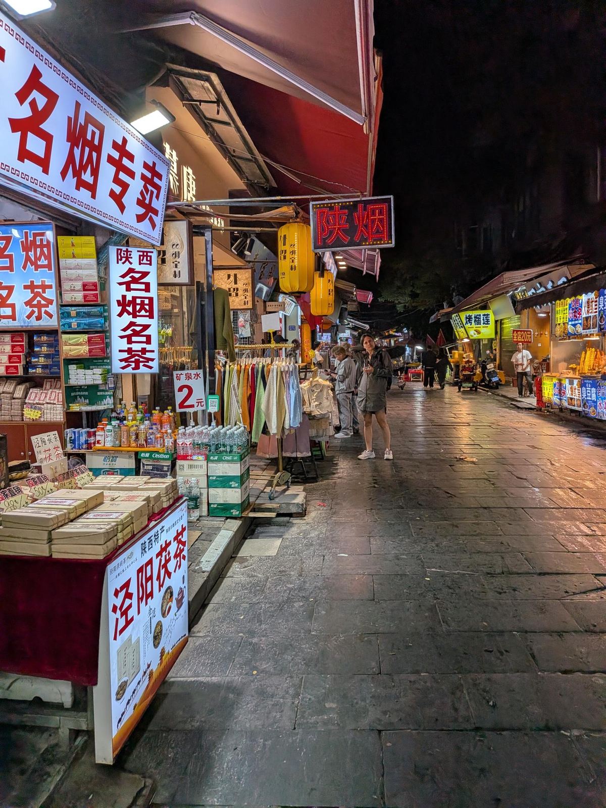Ruelle avec boutique de cigarettes et de thé (陕西名烟名茶), enseignes de street food et lanternes jaunes au-dessus du pavé mouillé
