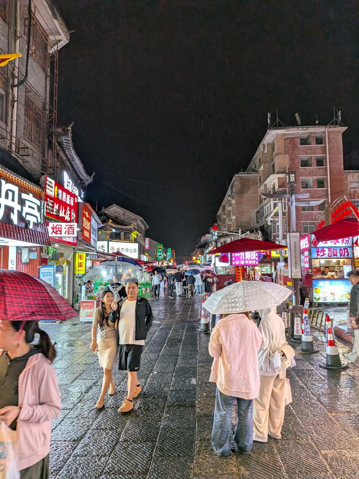 La rue piétonne sous la pluie, parapluies transparents et à carreaux, enseignes 烟酒超市, 毛蛋, 书亦烧仙草, stands de 炒酸奶 et 凉粉 sous les auvents rouges