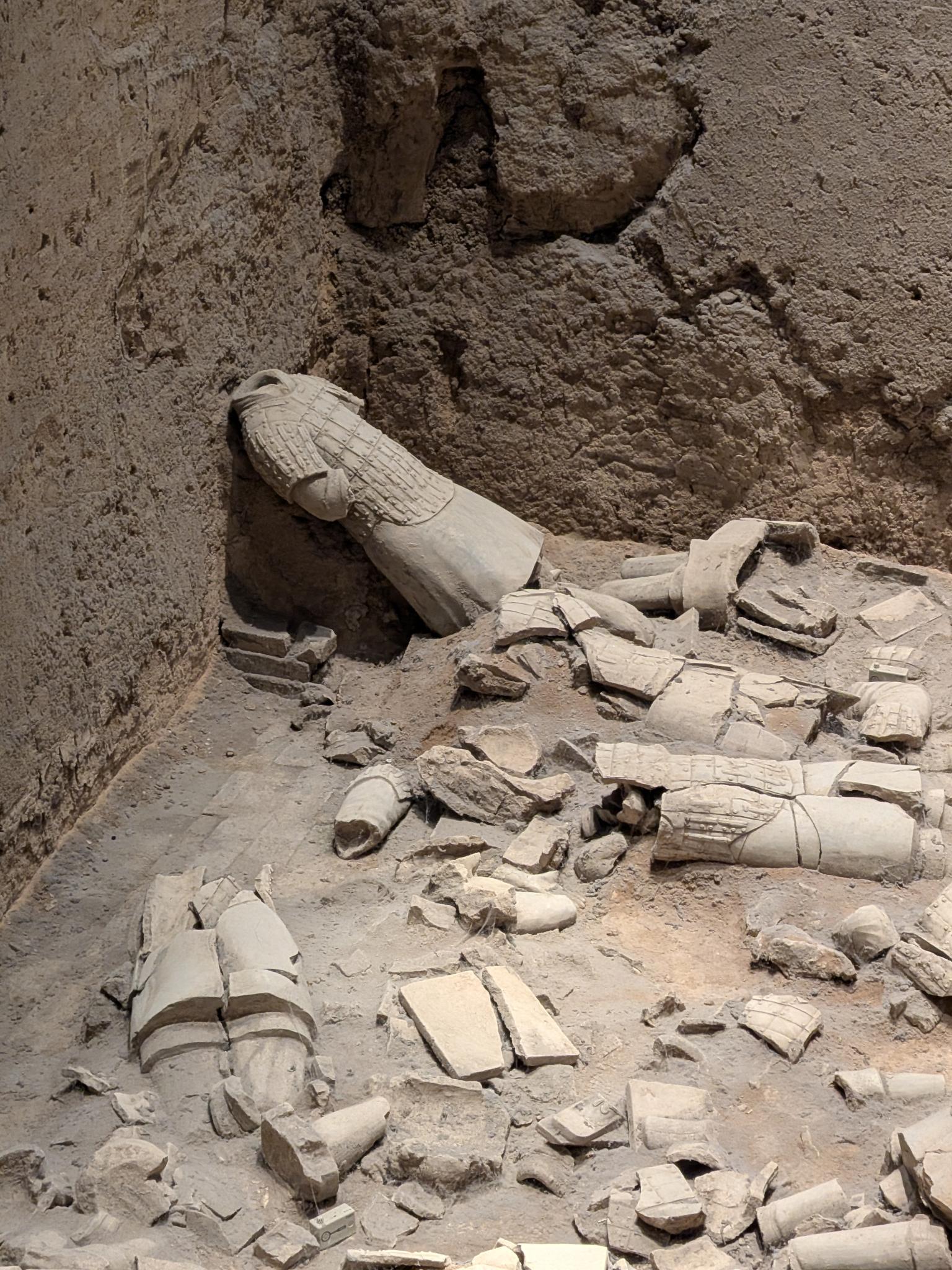 Guerrier adossé au mur de terre, armure à écailles fine, tête manquante, céramique brisée au sol