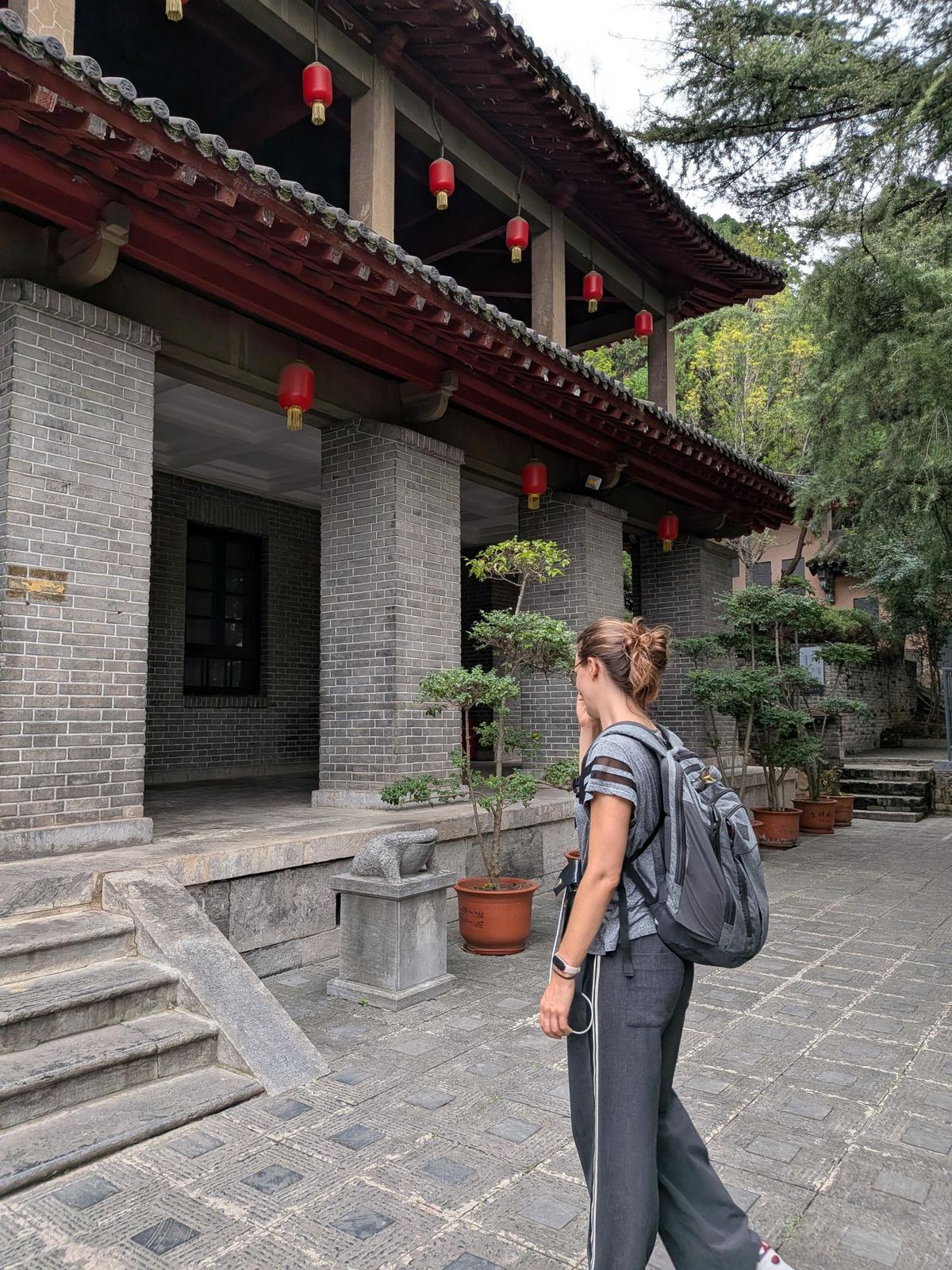 Marion qui traverse la cour pavée du temple, bâtiment en brique grise à colonnes, lanternes rouges et pots de saule en terre cuite le long du chemin