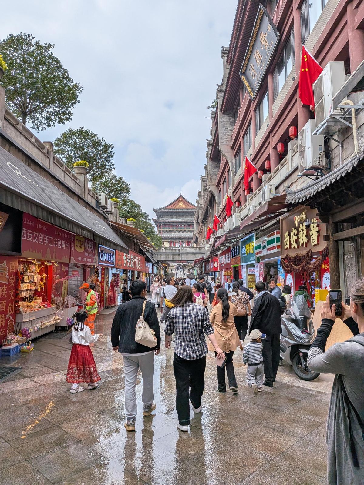 Rue commerçante sous la pluie : enseignes 西安文创手工非遗集市, 大唐汉服租赁, 长安旅行商店, tour de porte au bout de la rue