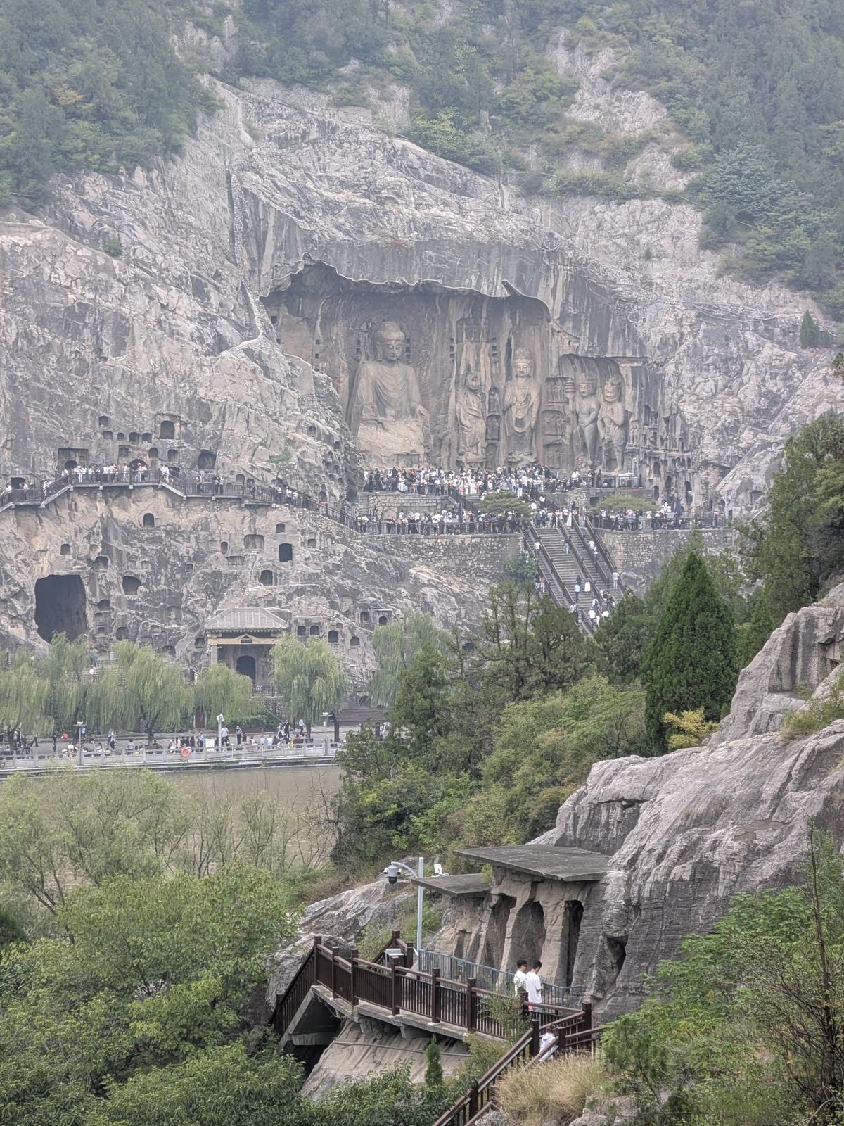 Le Bouddha de Fengxian et ses acolytes gravés dans la falaise, centaines de niches vides autour, passerelle en bois et petit pavillon en bord de rivière boueuse