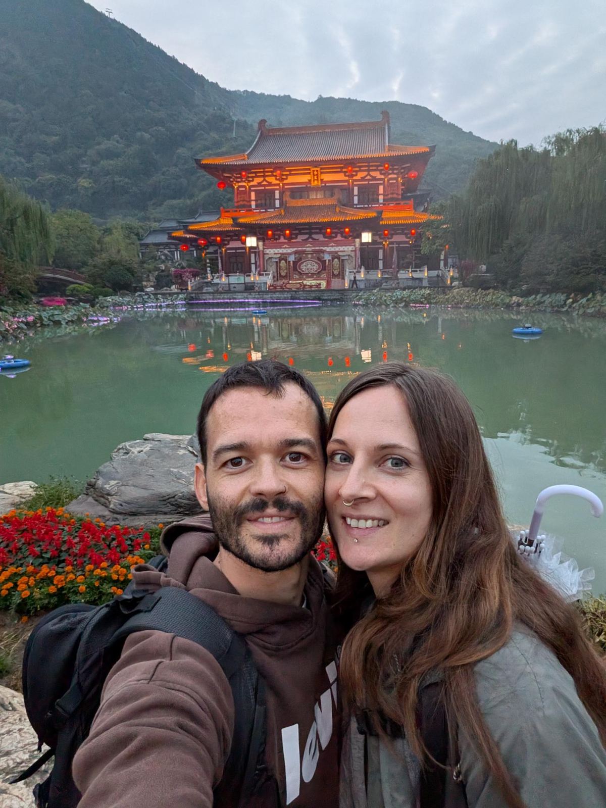 Selfie du couple devant le lac vert reflétant le palais illuminé d'orange et les lanternes rouges, collines boisées en arrière-plan