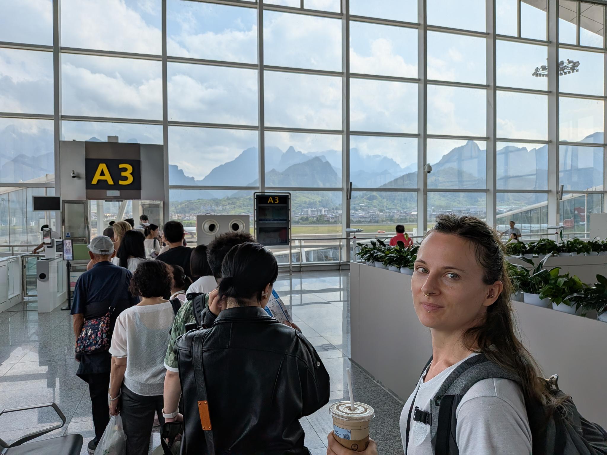 Marion au comptoir de check-in A3, son café glacé et les montagnes à travers la baie vitrée