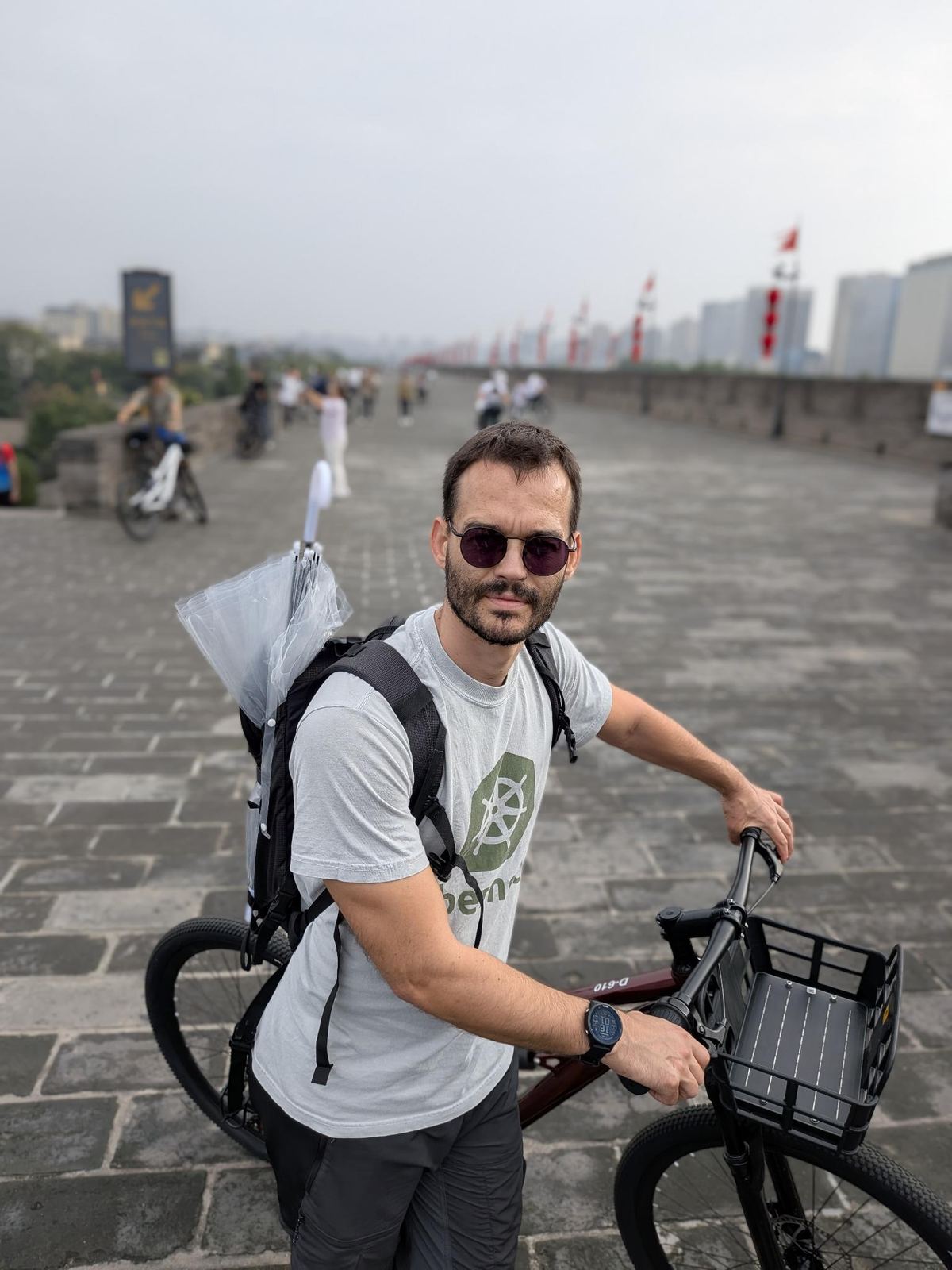 Tristan sur le rempart avec son vélo D-610 et son sac à dos, drapeaux rouges derrière lui, ville moderne au loin