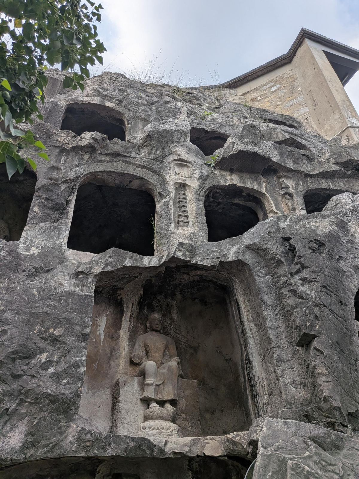 Le Bouddha assis sur son lotus érodé dans sa niche, petites stoupas et reliefs au-dessus, bâtiment traditionnel à toit tuilé derrière la falaise