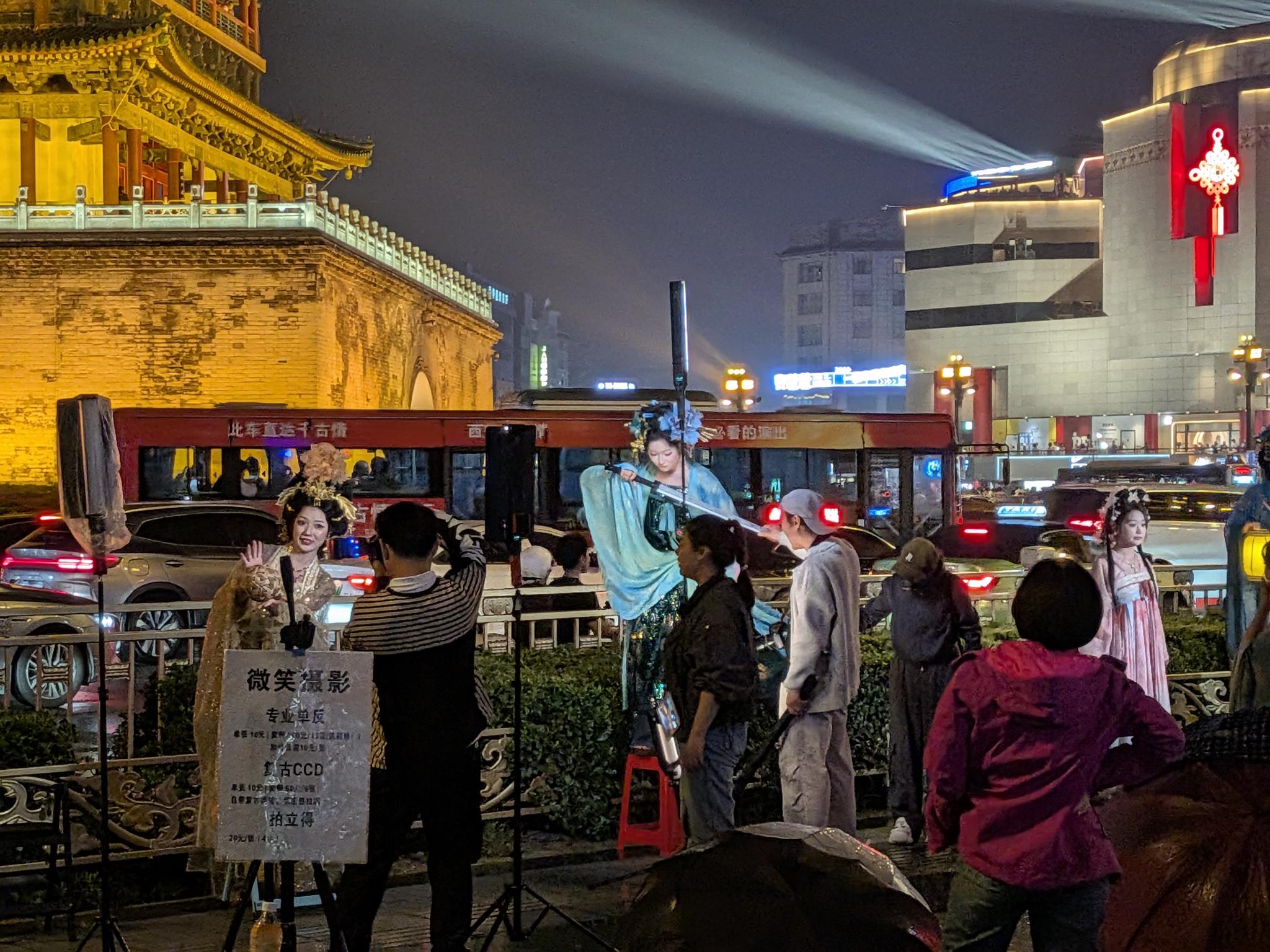 Photoshoot hanfu de nuit devant la porte illuminée, photographe au trépied et panneau 微笑摄影, bus touristique 千古情 en arrière-plan