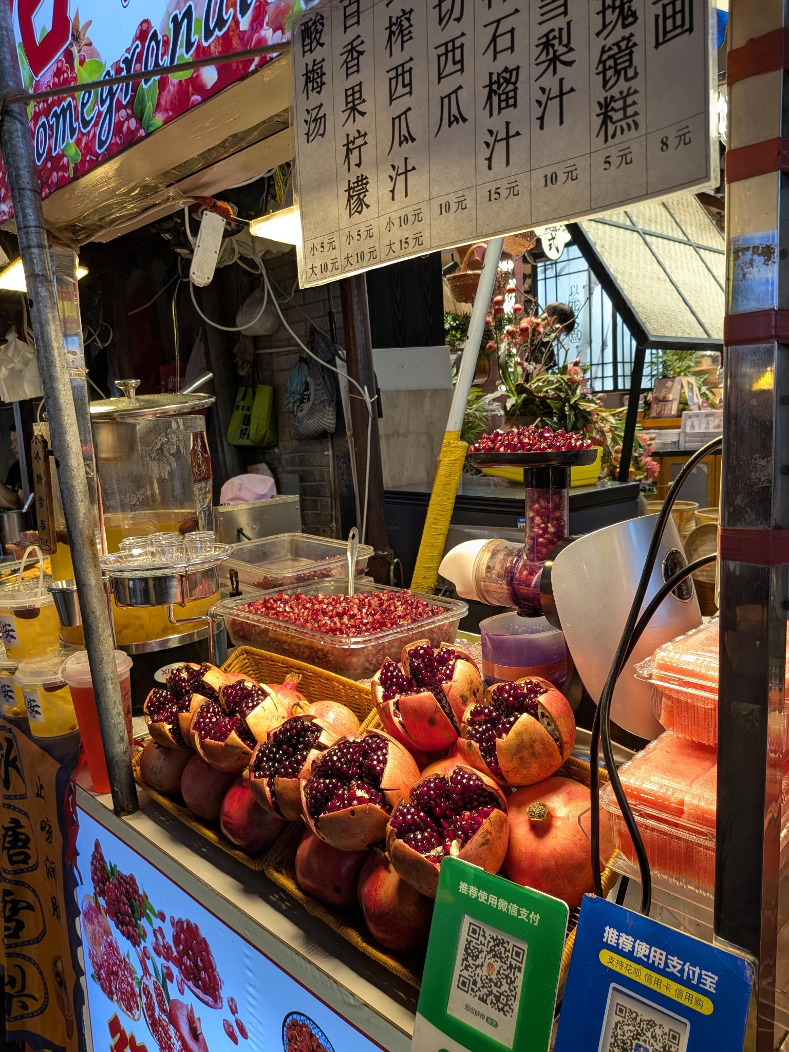 Stand de jus de grenade, grenades entières et coupées, presse manuelle, tableau des prix (石榴汁 15 ¥) et QR codes de paiement