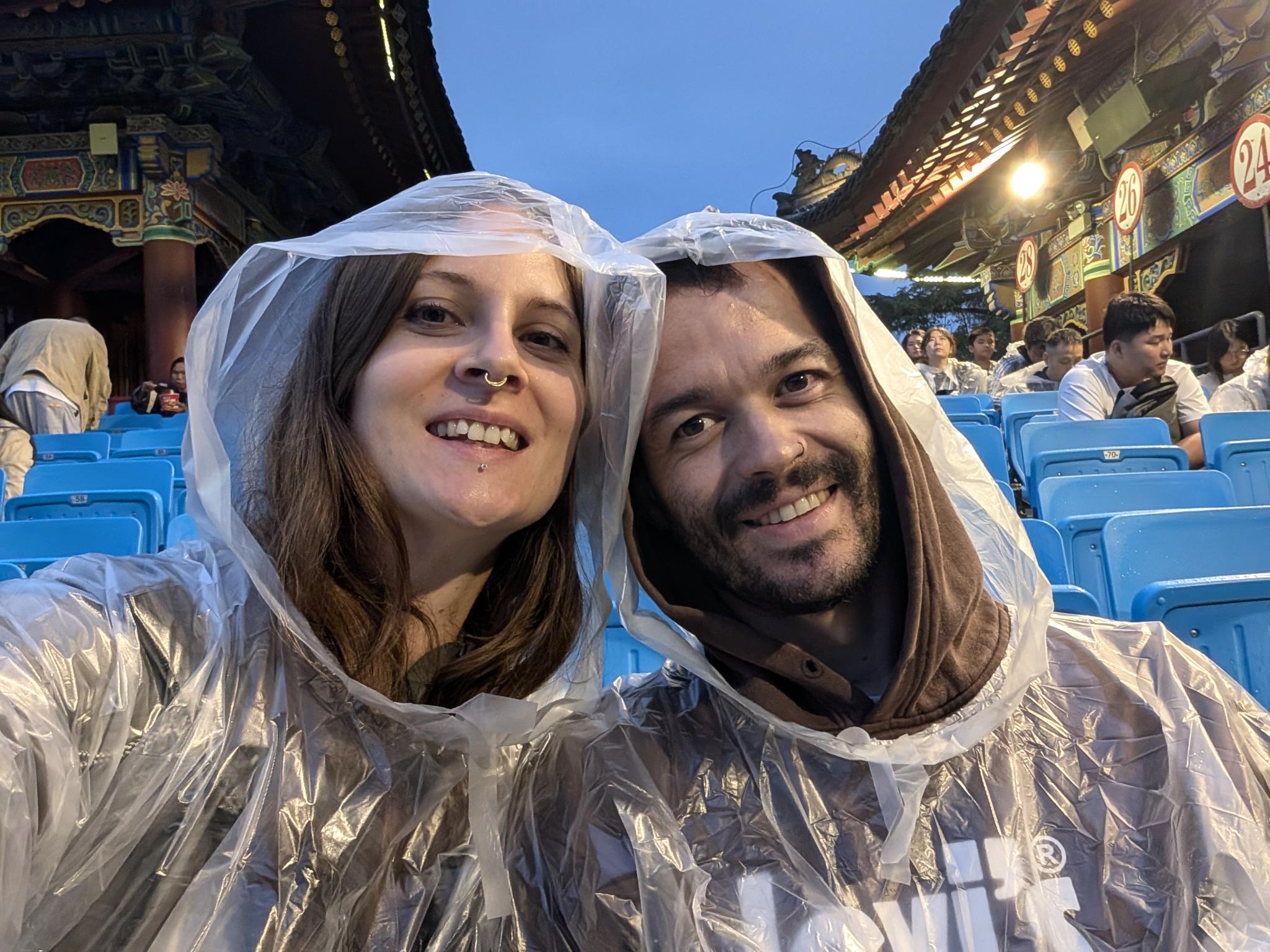 Selfie du couple en k-way transparent sur les gradins bleus, architecture traditionnelle lumineuse en arrière-plan