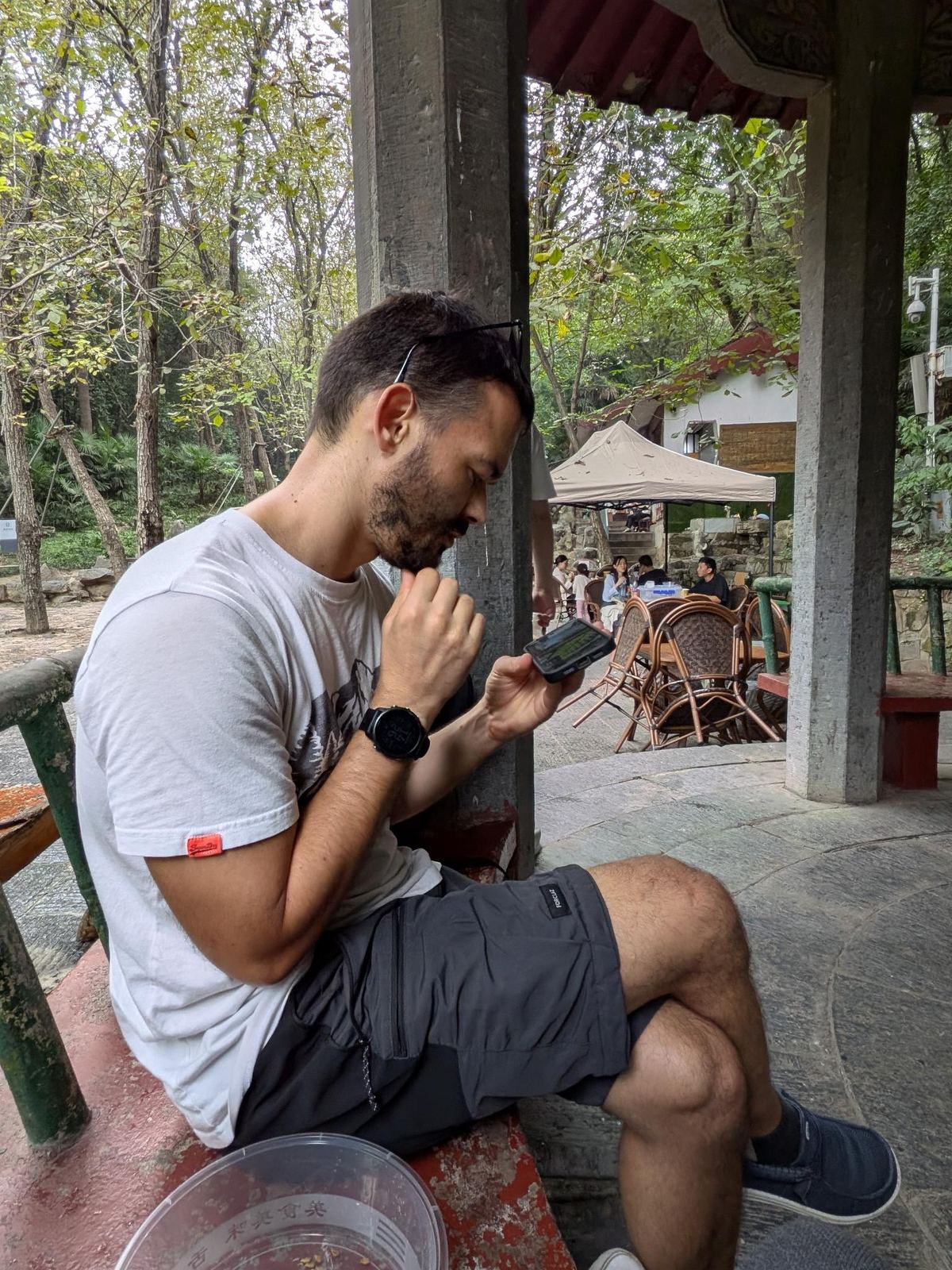 Tristan assis sur son banc rouge au pavillon, menton dans la main, son téléphone GPS à l'autre main, tentes et tables au loin dans le parc