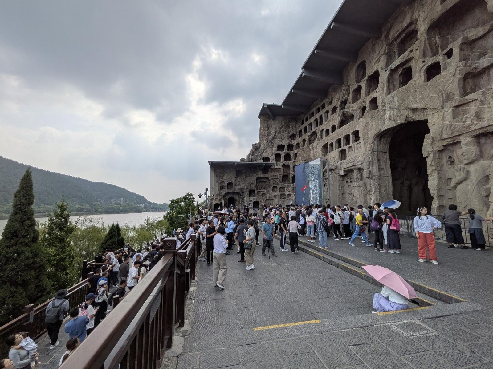 La place devant la grande grotte du Bouddha assis, foule de touristes aux parasols rose et bleu, parapet en bois en bord de rivière