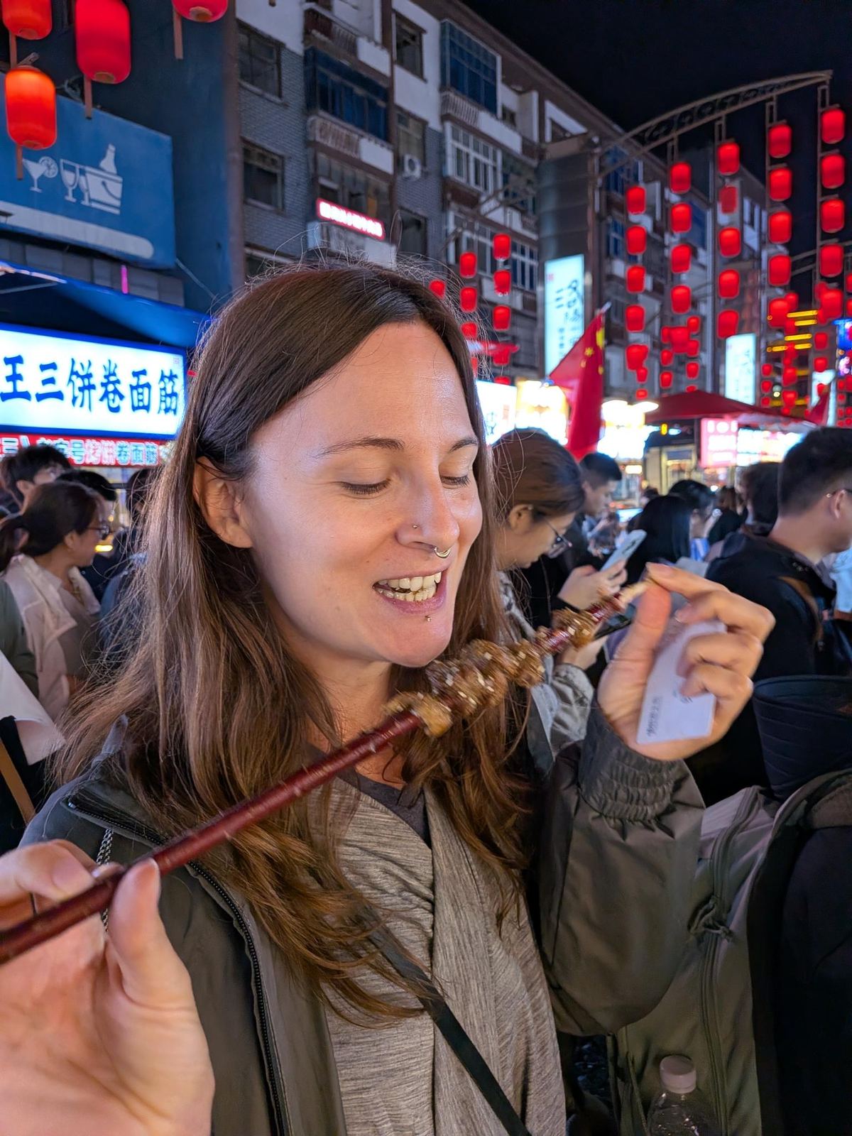 Marion qui mord dans sa brochette grilée, rangée de lanternes rouges au-dessus et enseigne 王三饼卷面筋 à gauche, foule en arrière-plan