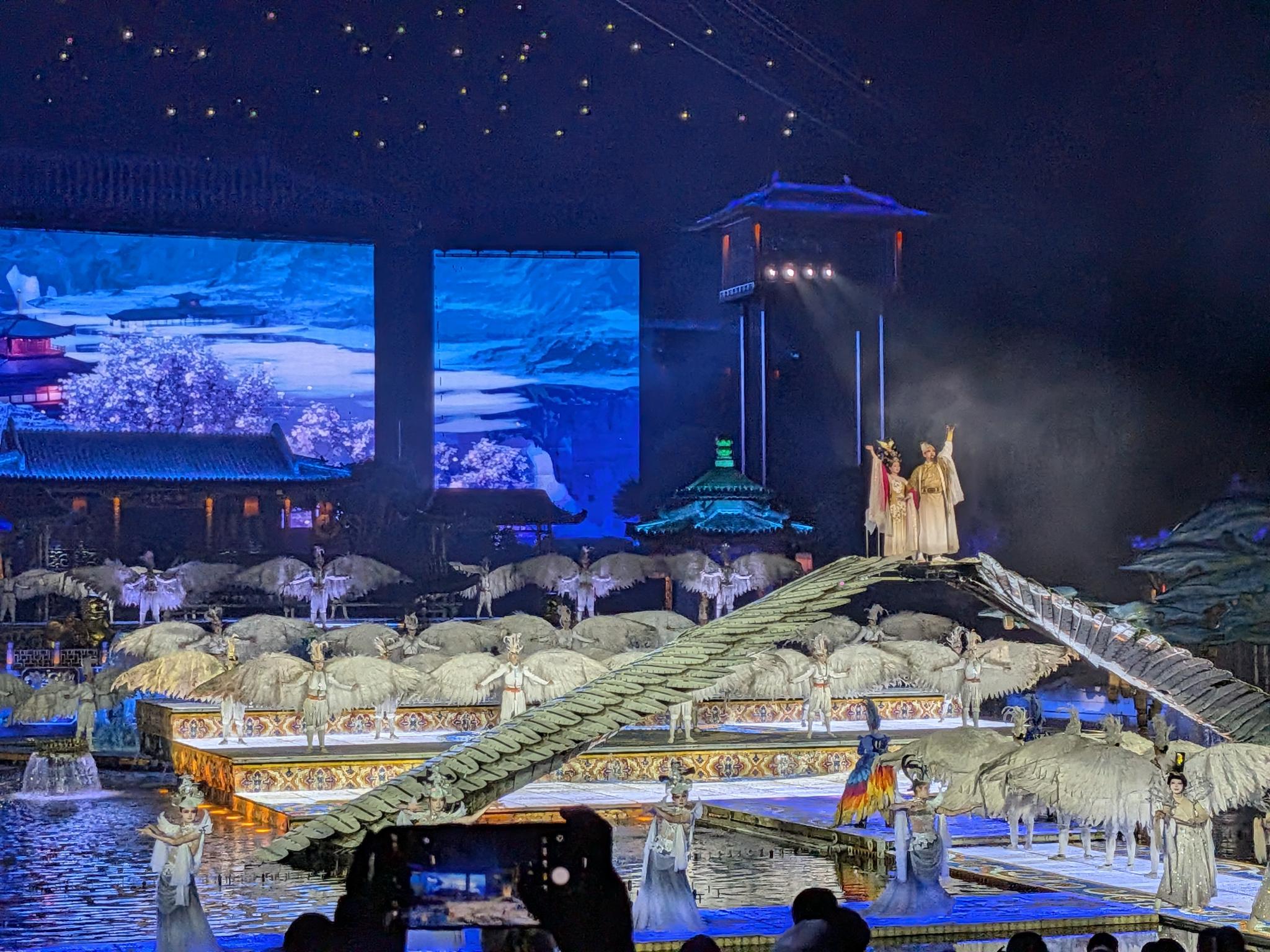 Danseuses en costumes blancs et ailes déployées sur les plateformes flottantes, deux acteurs sur le toit tuilé, écrans de montagne et pavillon illuminé