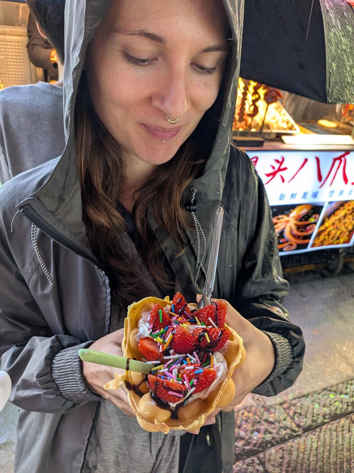 Marion qui tient sa gaufre façon Hong Kong : cône gaufre, glace, fraises, confettis et chocolat, stand de rue sous auvent noir et pavés mouillés