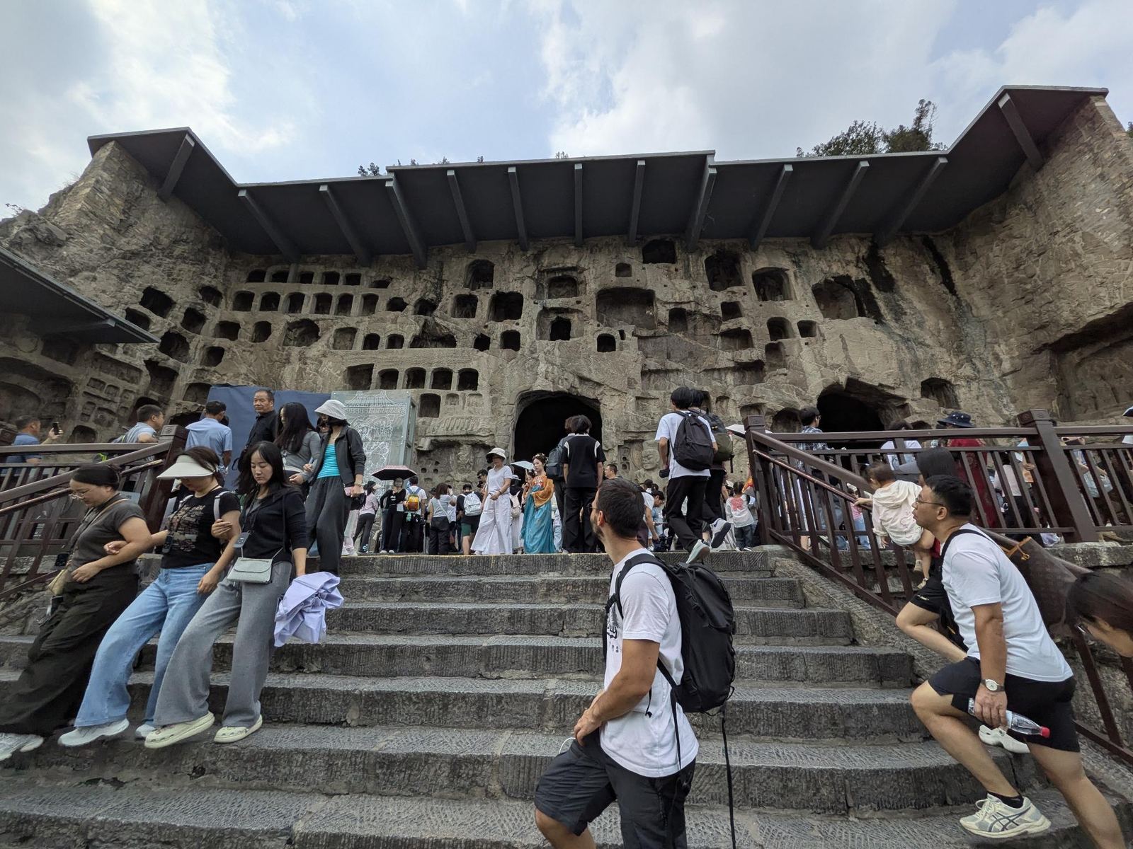 La falaise de niches sous son toit de protection moderne, escalier en pierre au premier plan et touristes en remontée