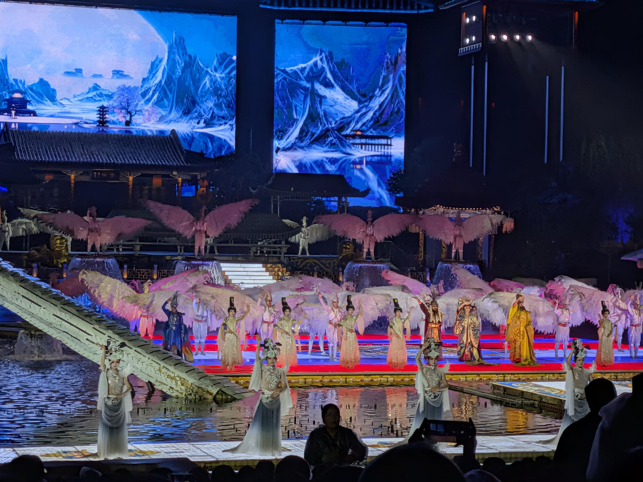 Scène sur l'eau avec des dizaines de danseuses à ailes roses et blanches, silhouettes de spectateurs et grands écrans LED bleus en arrière-plan