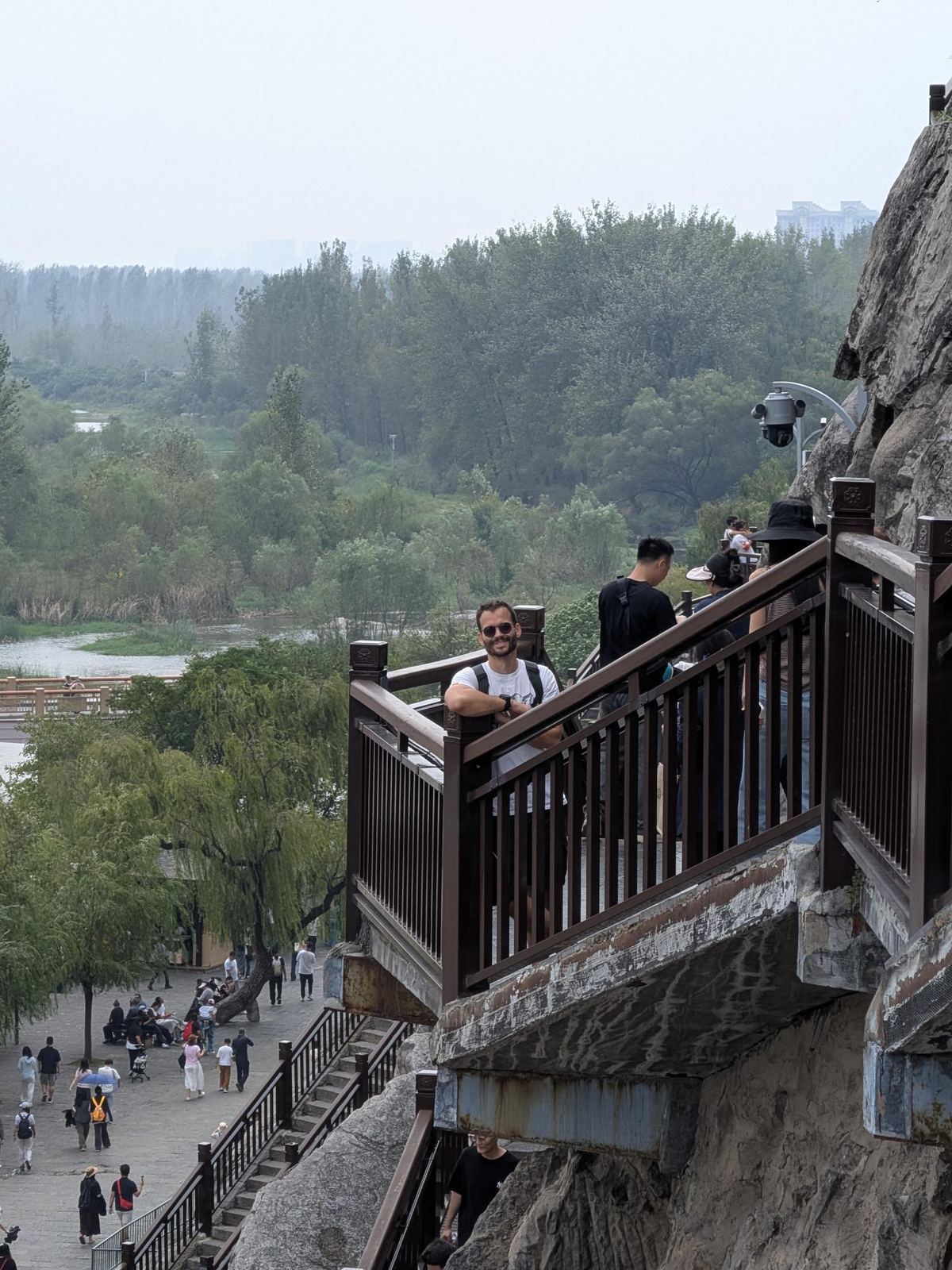 Tristan penché au parapet de la passerelle dans la falaise, rivière, saules et ville lointaine en contrebas, ciel gris
