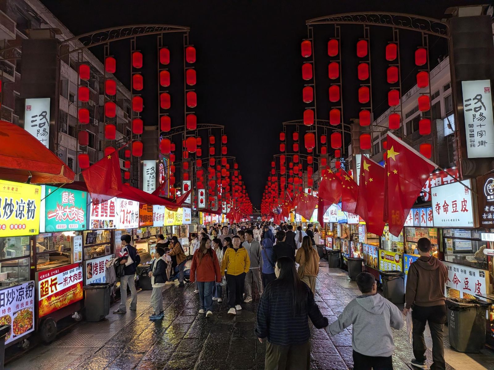 La rue de marché de Luoyang la nuit, enseignes 洛阳十字街, arches de lanternes rouges, drapeaux nationaux et stands illuminés (炒凉粉, 超长一米大肉串, 烤鳄鱼, 门钉肉饼), pavé mouillé et foule dense