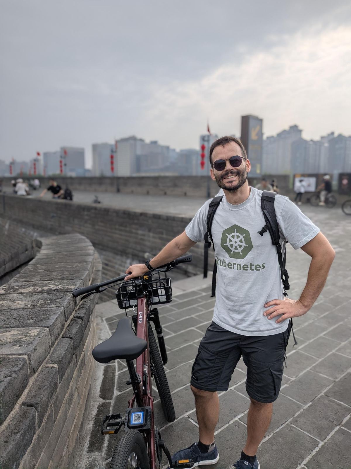 Tristan souriant en tee Kubernetes, adossé au vélo D-610 sur le rempart, drapeaux rouges et immeubles au loin