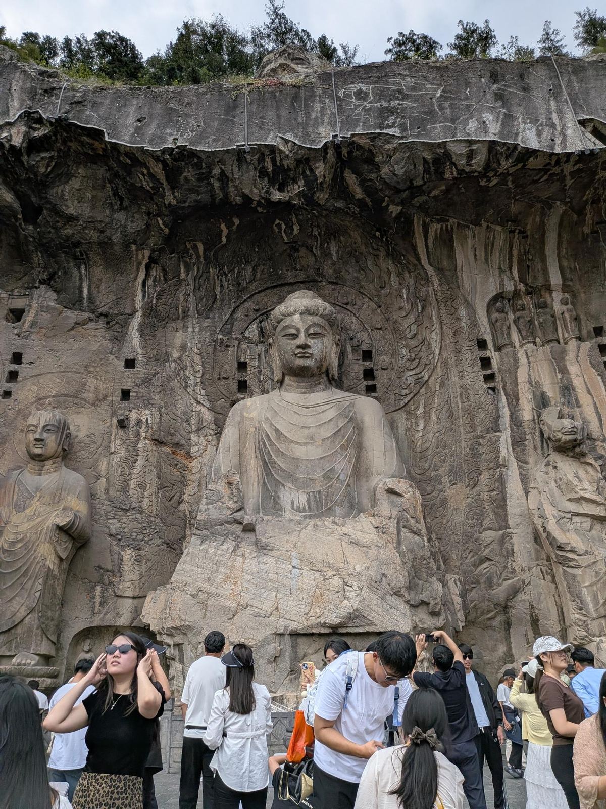 Le Bouddha assis vu de face, statues de cour de chaque côté et rangées de petites niches de Bouddha sur la falaise érodée