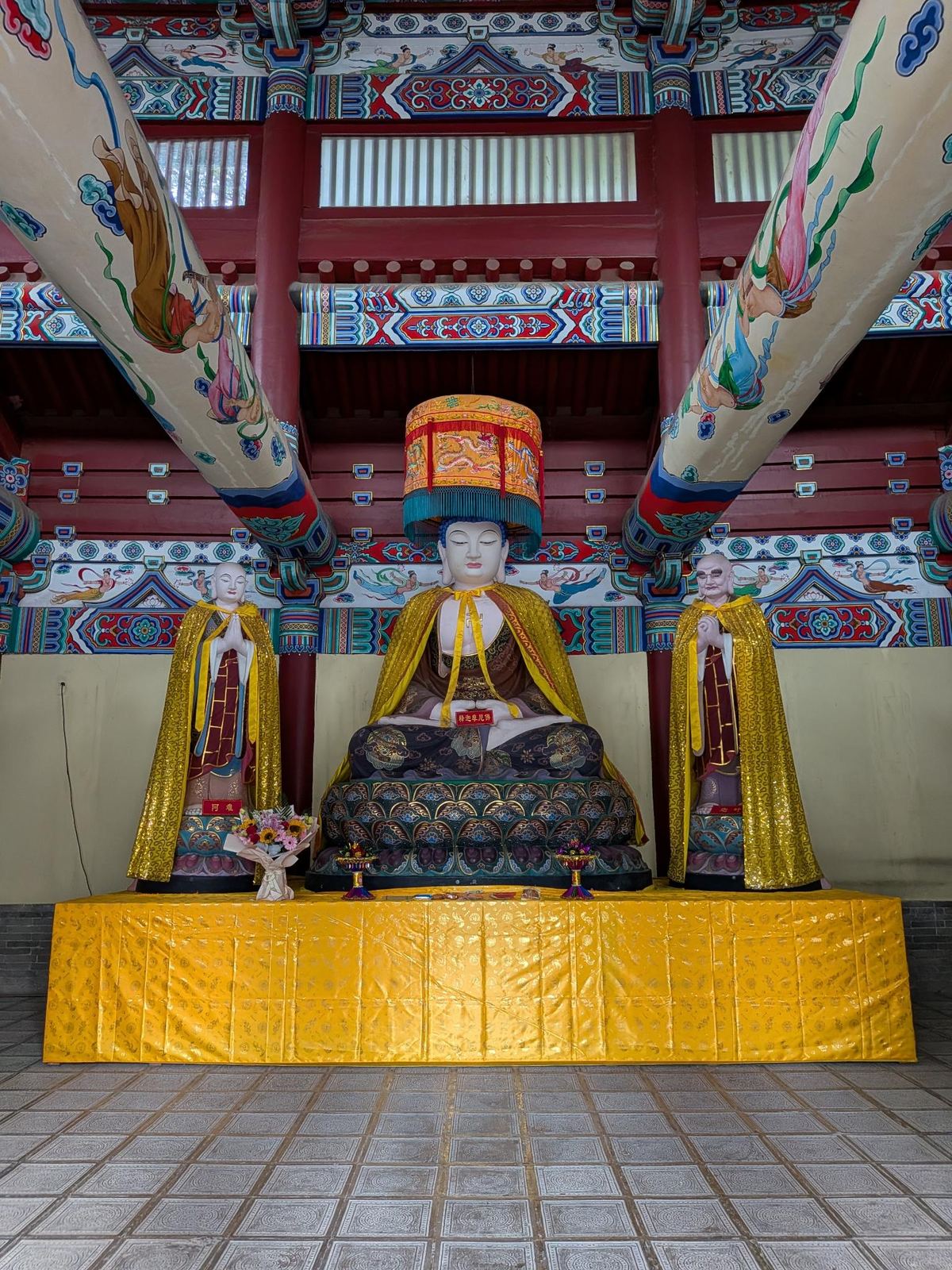 Le Bouddha assis doré sur son trône de lotus, auréole et canopée rouge, deux acolytes debout aux mains jointes, étiquettes 释迦牟尼佛 et 阿难