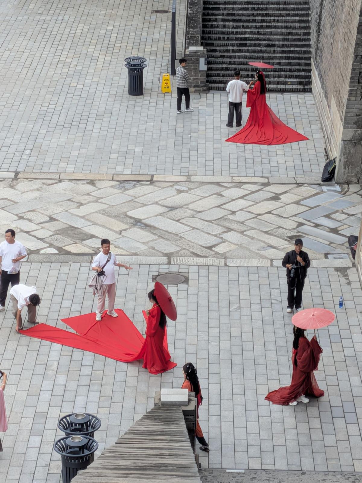Photoshoot hanfu sur la place devant la porte : mannequins en robes rouges à traîne, parasols rouges, assistants et photographe
