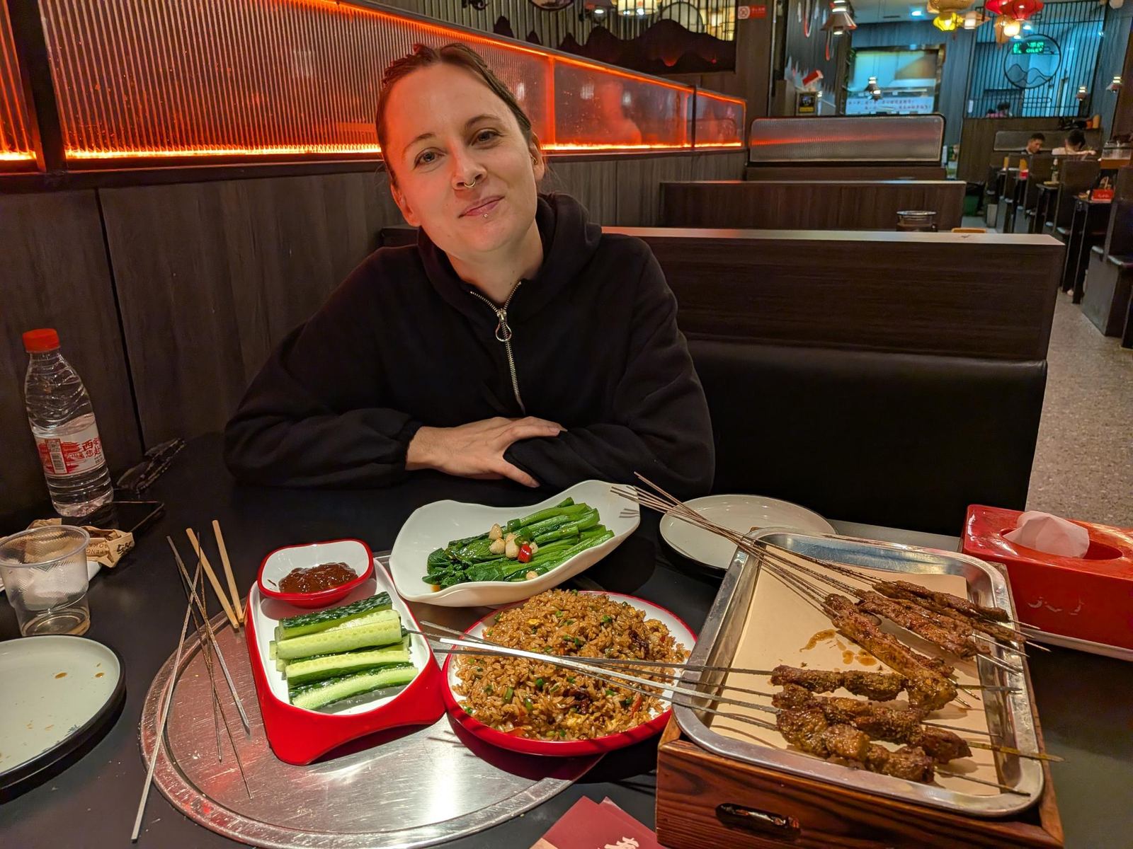 Marion en pull zippé noir, bras croisés devant sa table, plateau de brochettes, riz sauté, légumes sautés et petits pois, bol de sauce, bouteille d'eau au logo rouge