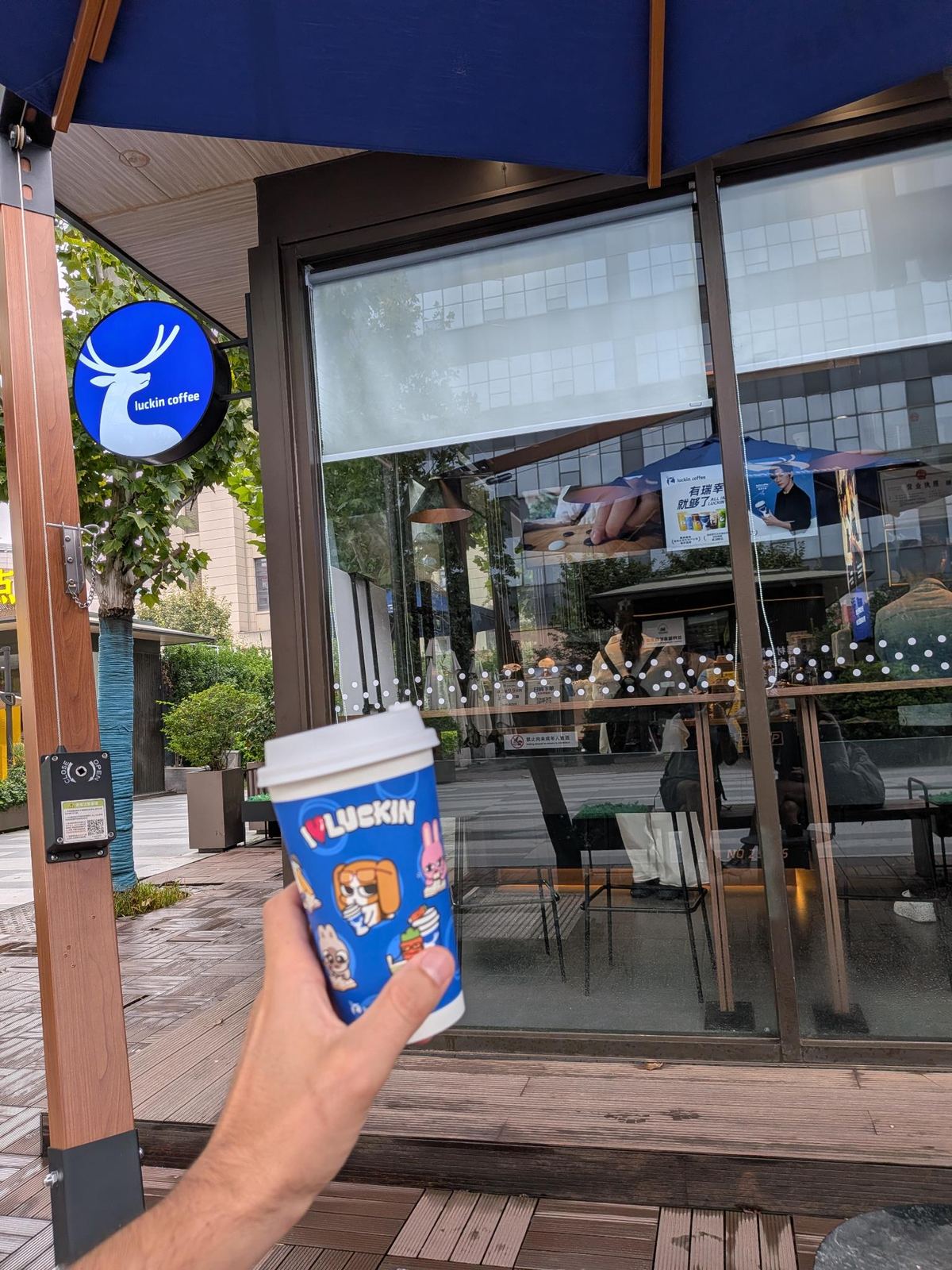 Tasse bleue Luckin Coffee avec mascottes cartoon devant la boutique Luckin, terrasse en bois sous ombrelle bleue