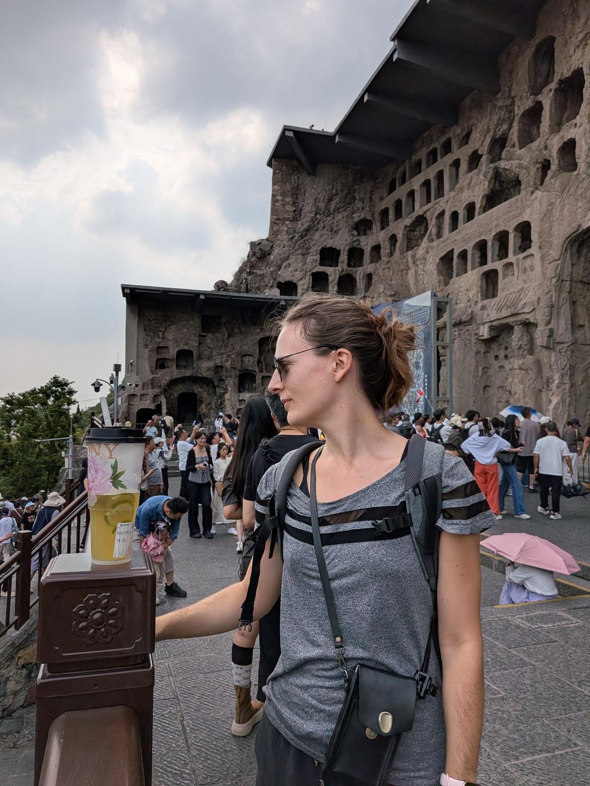 Marion de profil devant le parapet sculpté, sa boisson glacée à la fleur rose devant elle, foule et falaise de niches en arrière-plan