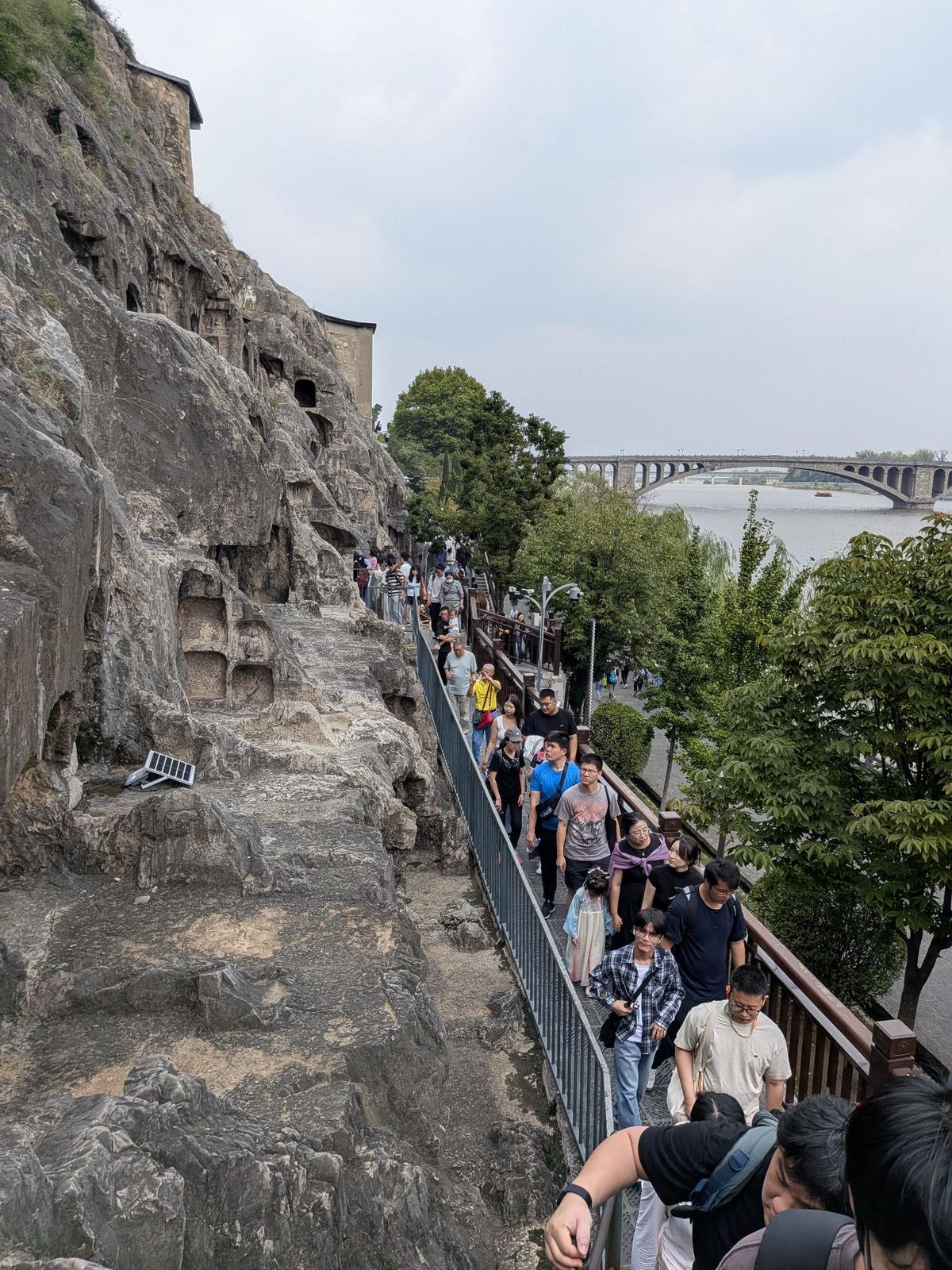 La passerelle au pied de la falaise, file de touristes, rivière et grand pont en arc au loin sous le ciel gris