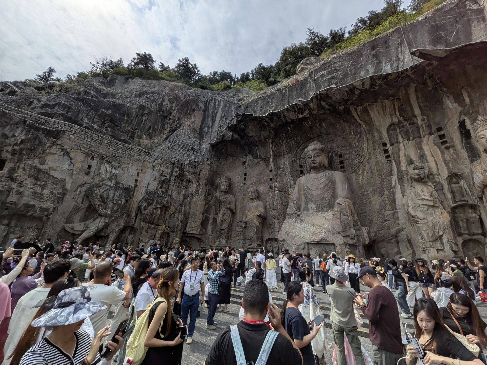 Vue plongeante sur la grande grotte et le Bouddha assis, falaise entièrement sculptée, filet de protection au sommet, foule sur l'esplanade