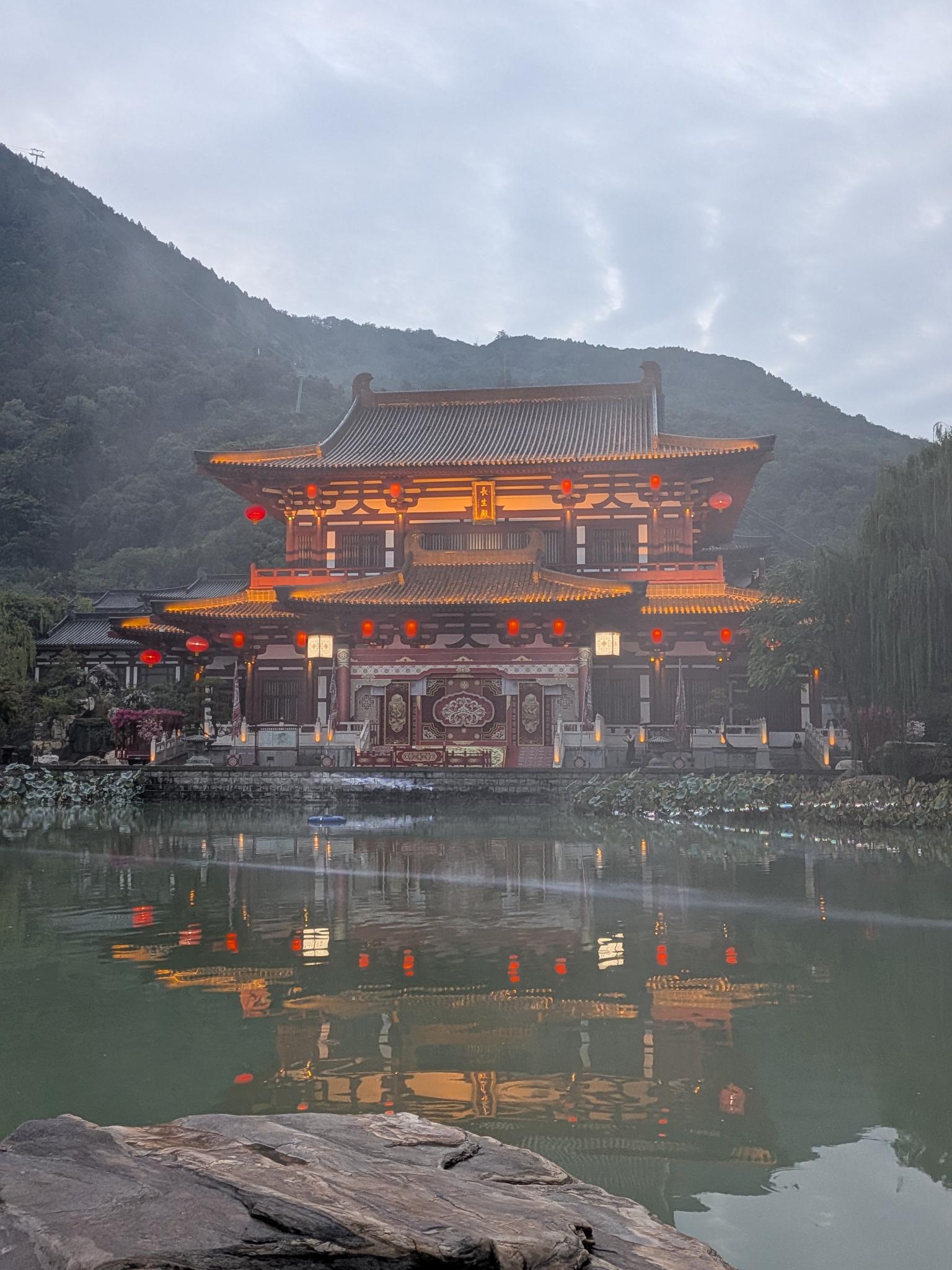 La salle 长生殿 (Changsheng, Hall de la Longue Vie) illuminée de nuit au bord de la piscine, lanternes rouges et collines boisées en arrière-plan