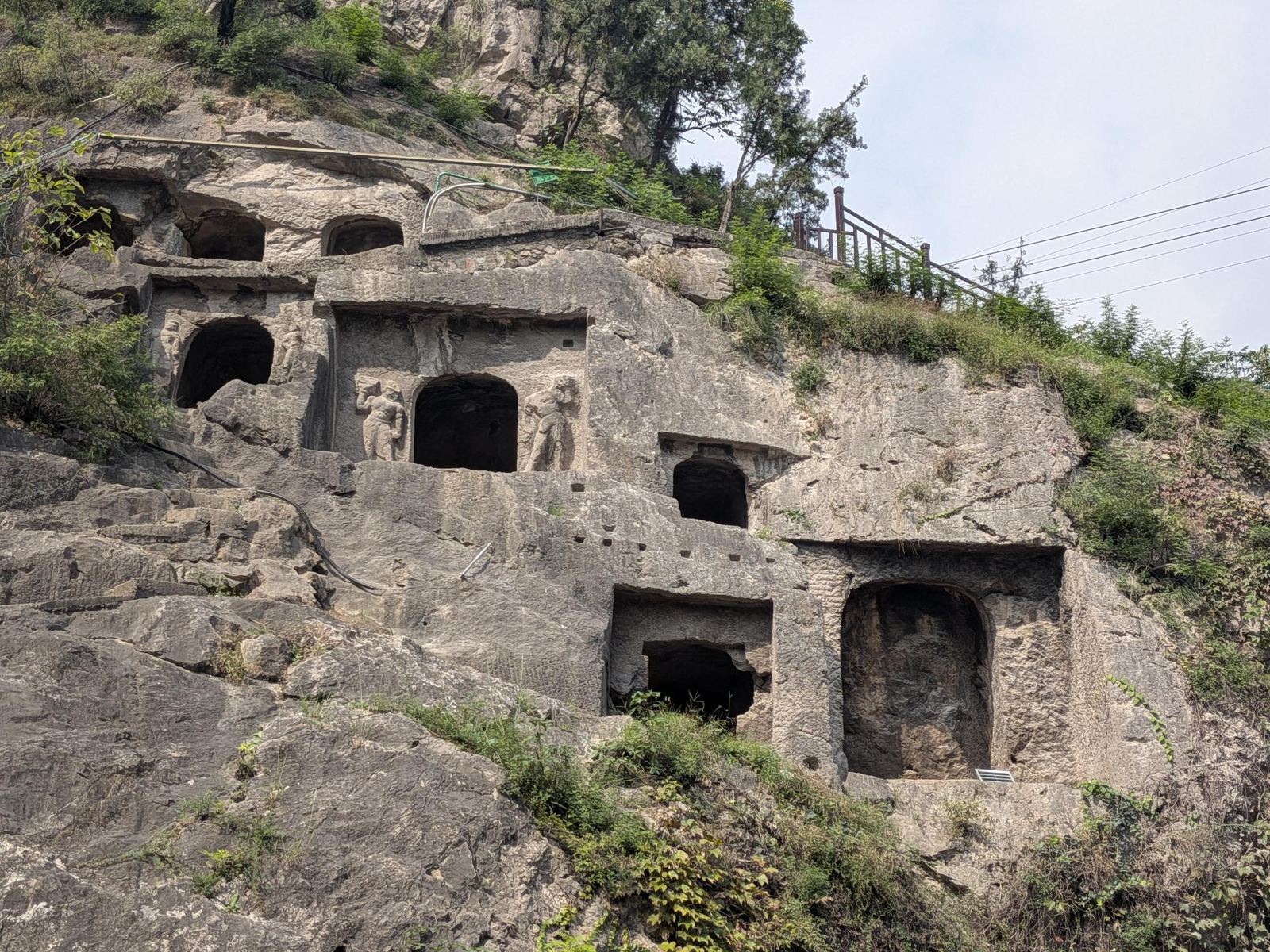 Deux statues debout encadrant une porte en arche dans la falaise sculptée, buissons verts au sommet et câbles au loin