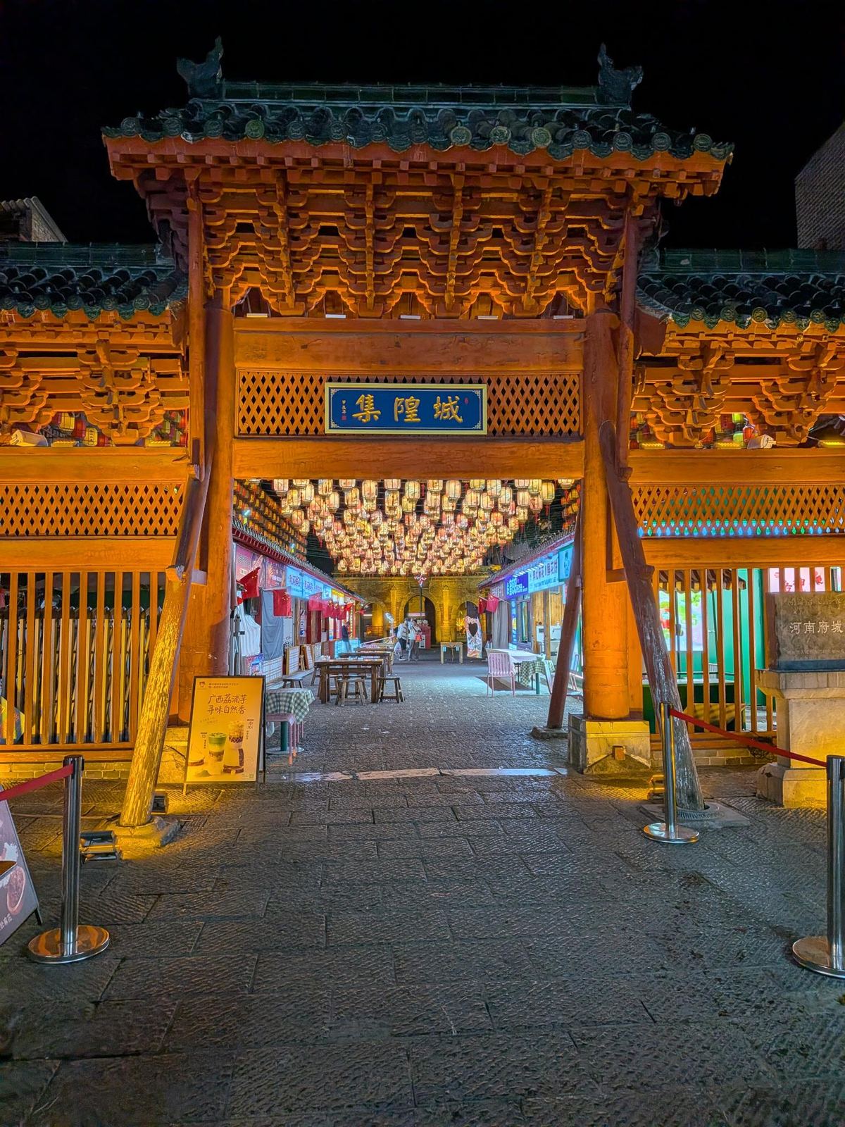 Porte triomphale traditionnelle illuminée d'orange la nuit, fronton 城隍集, stèle 河南府, rue pavée bordée de lanternes lumineuses et de boutiques au fond