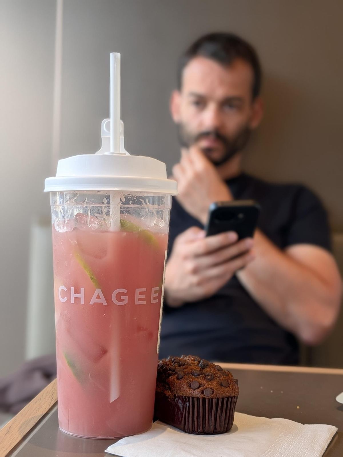 Gobelet de thé glacé rose CHAGEE au citron vert à côté d'un muffin au chocolat, Tristan au téléphone en arrière-plan flou