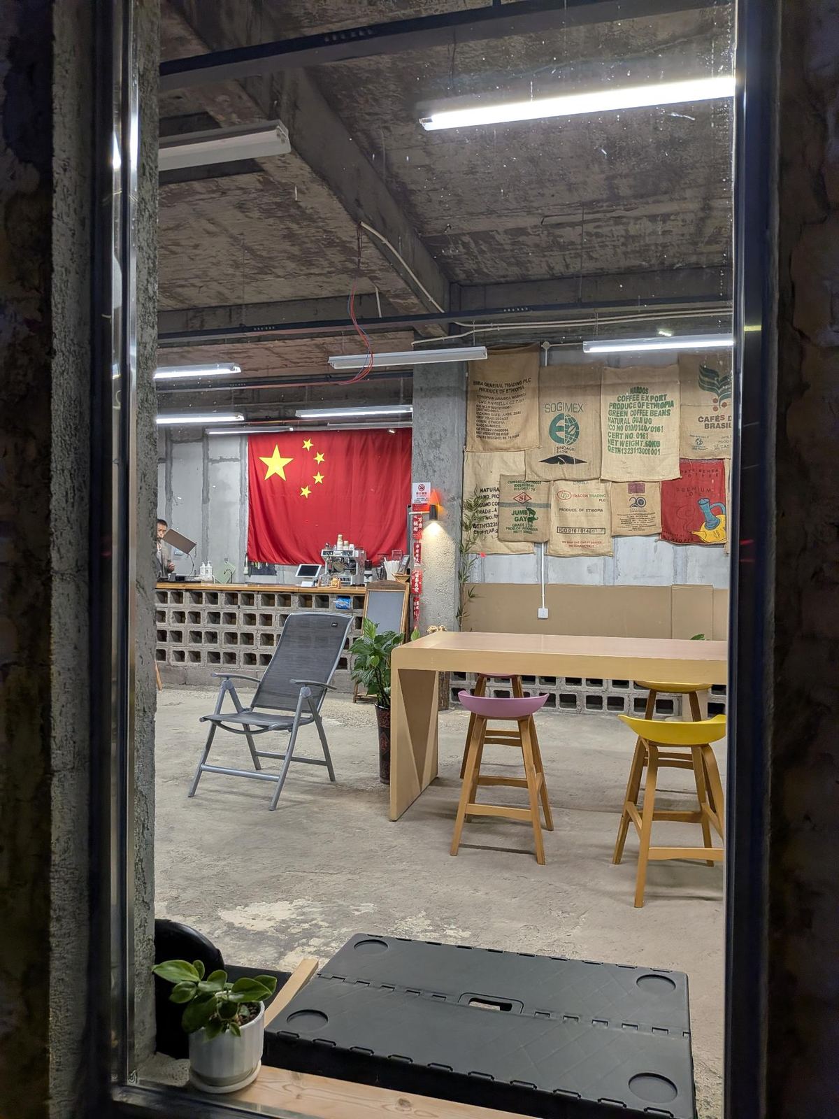 Café industriel vu de la rue, plafond béton et néons, drapeau national au mur, comptoir blocs de béton, sacs de café vintage ETHIOPIA et BRASIL, tabourets roses et jaunes