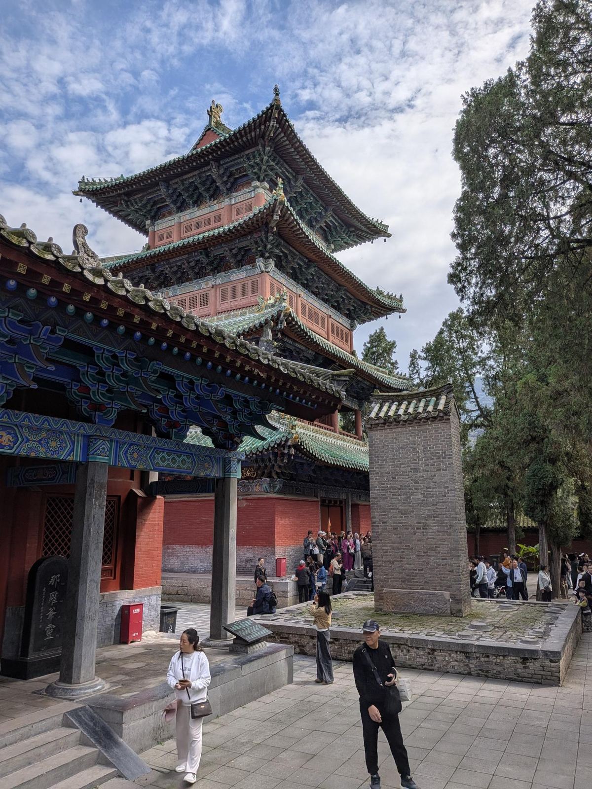 Pavillon à toits superposés vu de son couloir voûté, colonnes de pierre, stèle noire et piliers dans la cour, cyprès et ciel bleu