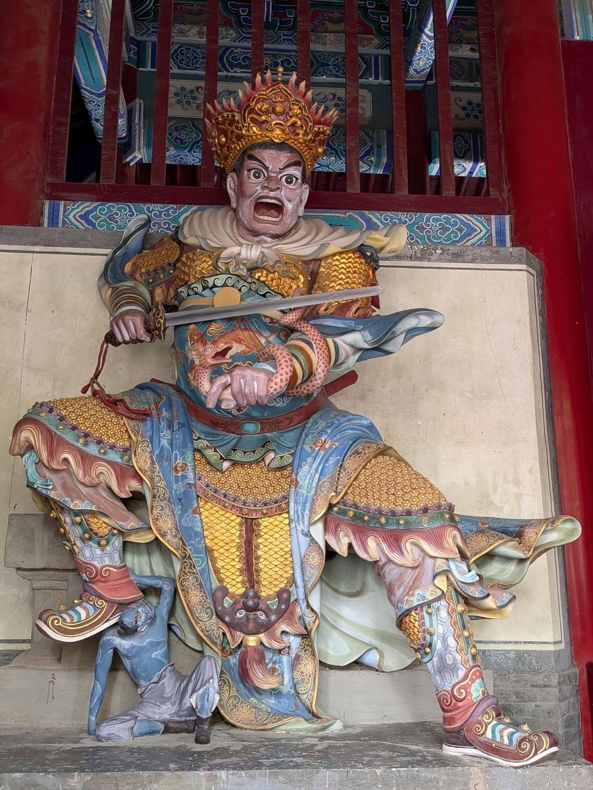 Statue peinte d'un gardien féroce au casque doré, armure, épée et dragon enlacé au bras, un pied sur un démon, devant les colonnes rouges du temple