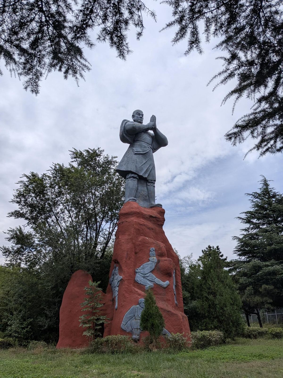Grande statue de bronze d'un moine de Shaolin, mains levées en salut, sur son socle de roche rouge orné de bas-reliefs de kung-fu, parc et conifères, ciel couvert