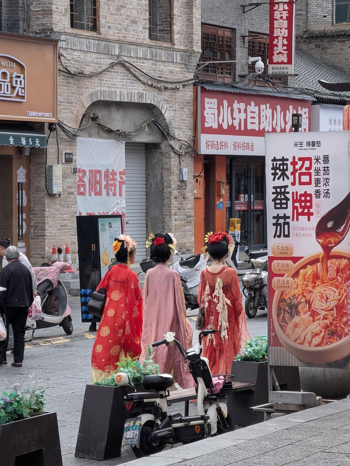 Rue commerçante de la vieille ville de Luoyang (南大街), trois femmes en hanfu rouge et rose, enseignes de hot pot et de produits locaux, scooter et bâtiments en brique