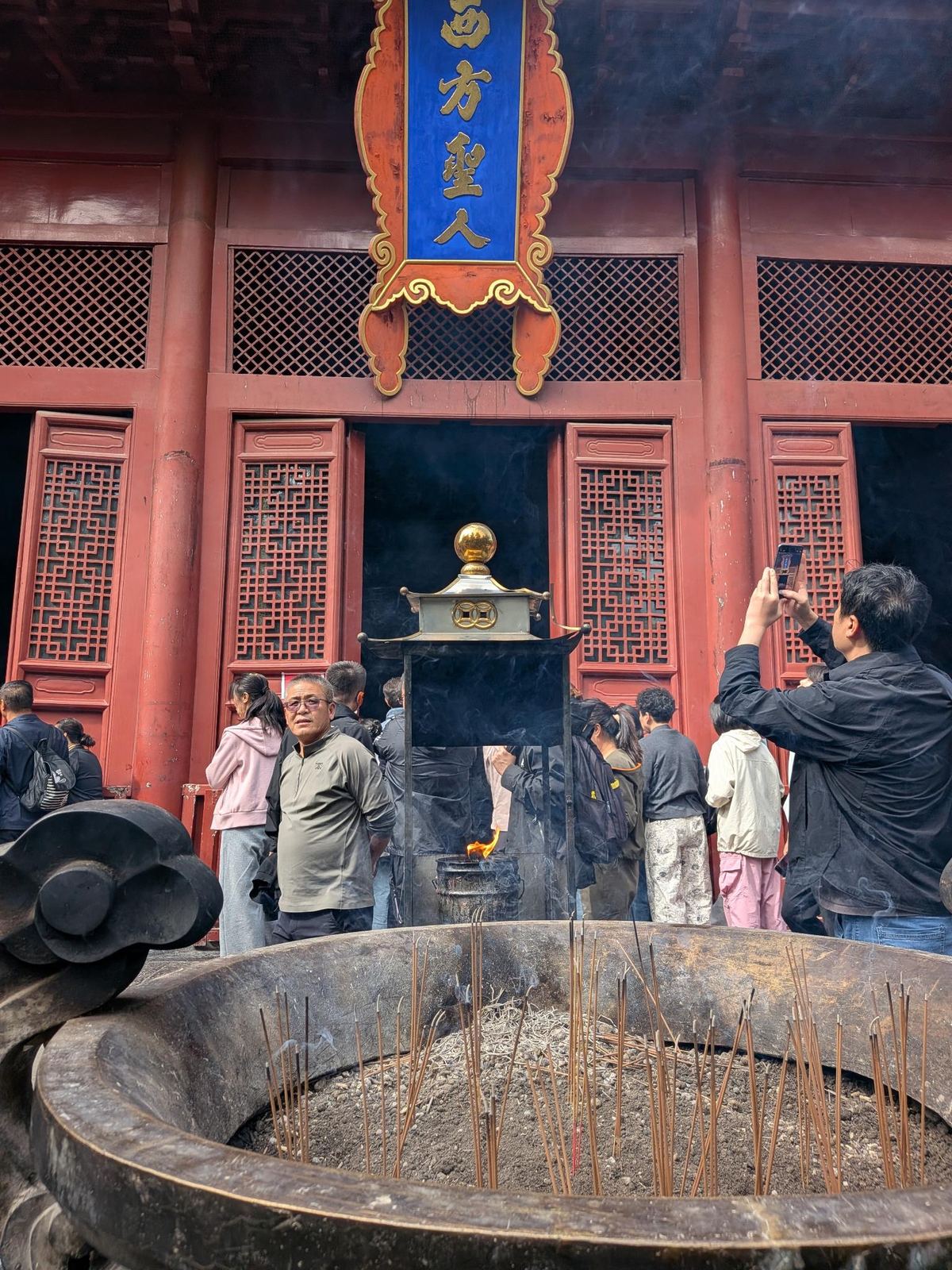 Encensoir en pierre plein de bâtonnets dressés, brûloir métallique qui fume derrière, foule de fidèles et touristes sous le fronton bleu 西方聖人