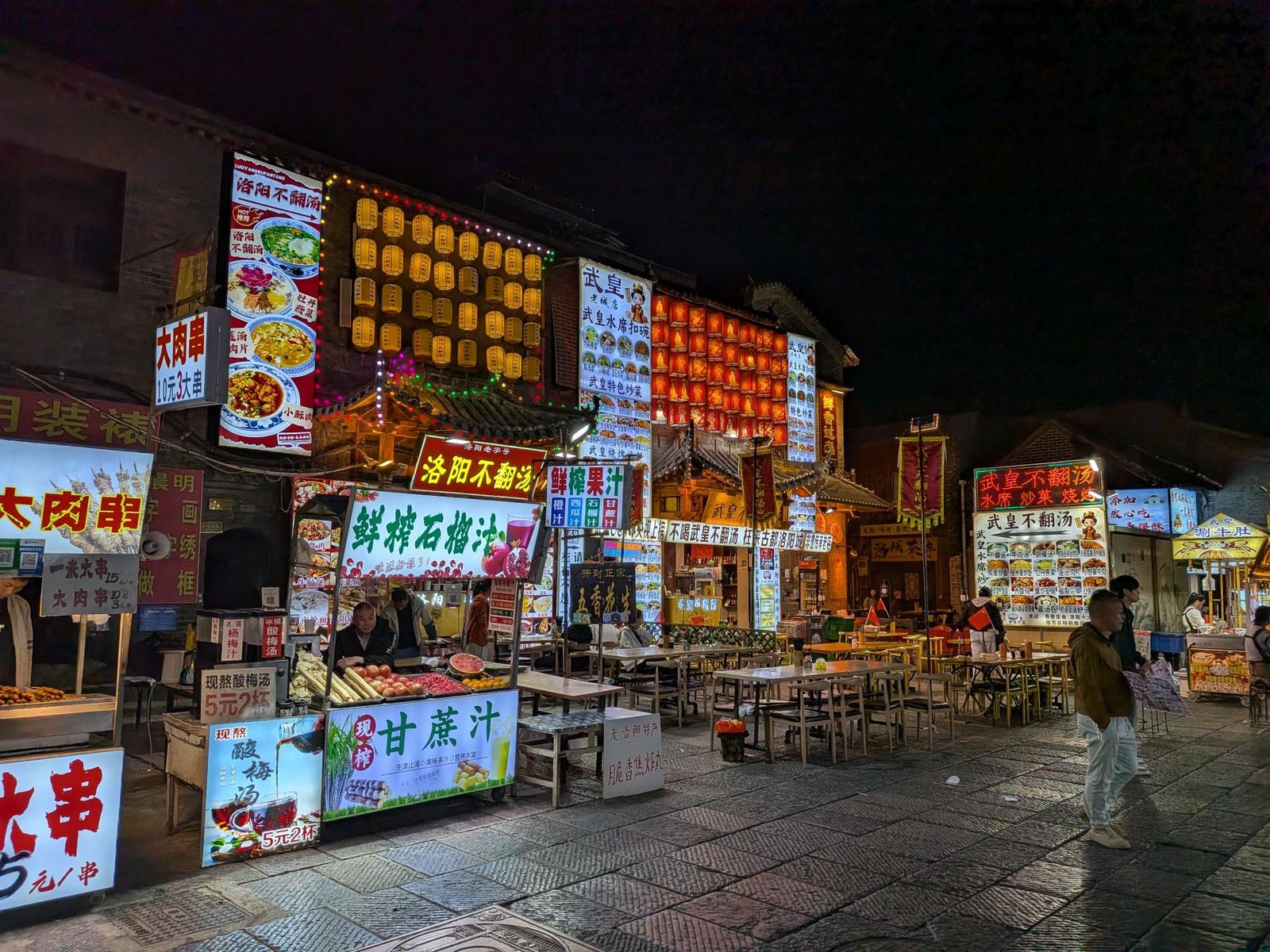 Rue de stands de rue illuminée la nuit, rangées de lanternes jaunes et rouges, enseignes 不翻汤, brochettes de viande, jus de grenade et de canne, tables dehors sur la place pavée