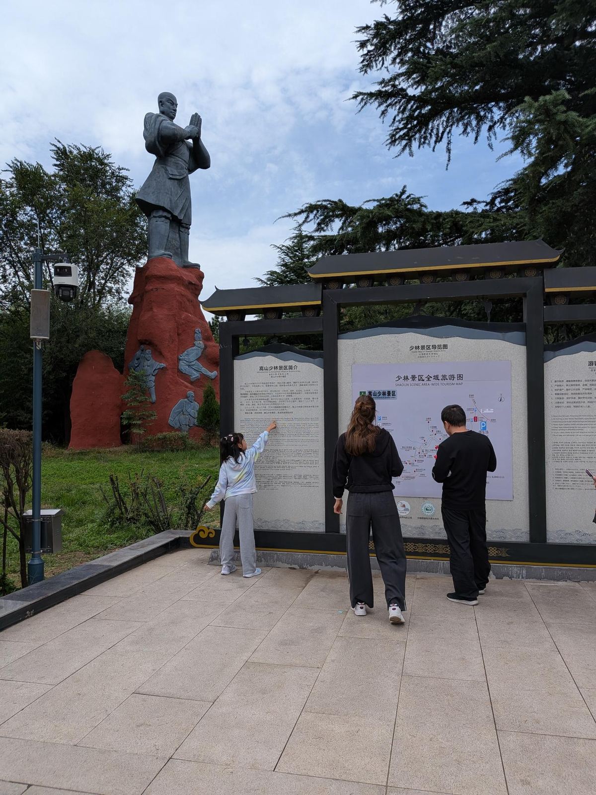La statue de moine de l'entrée et les panneaux de la carte du parc 少林景区全域旅游图, enfants et panneaux d'information sous un toit traditionnel