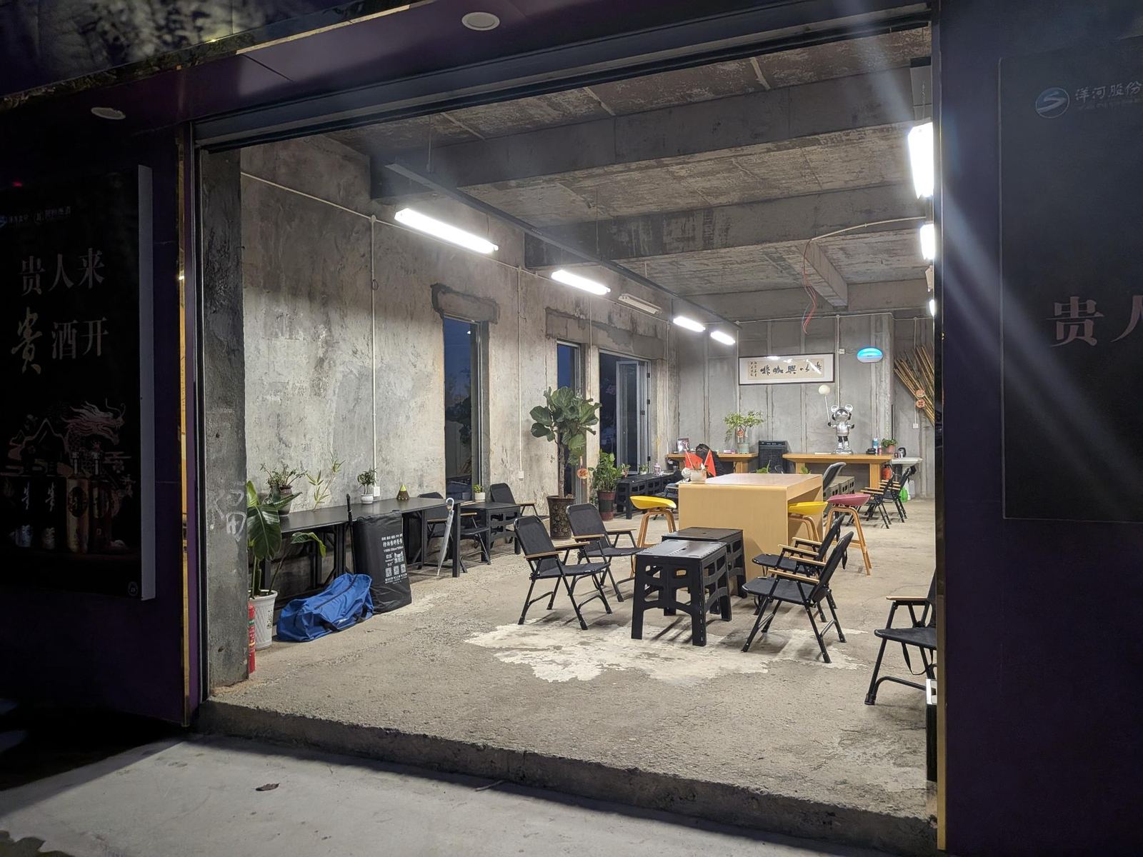 Intérieur du café, murs et plafond béton, néons en rangées, tables et chaises pliantes, plantes, poster 贵人来 酒开 et logo 洋河股份 au mur