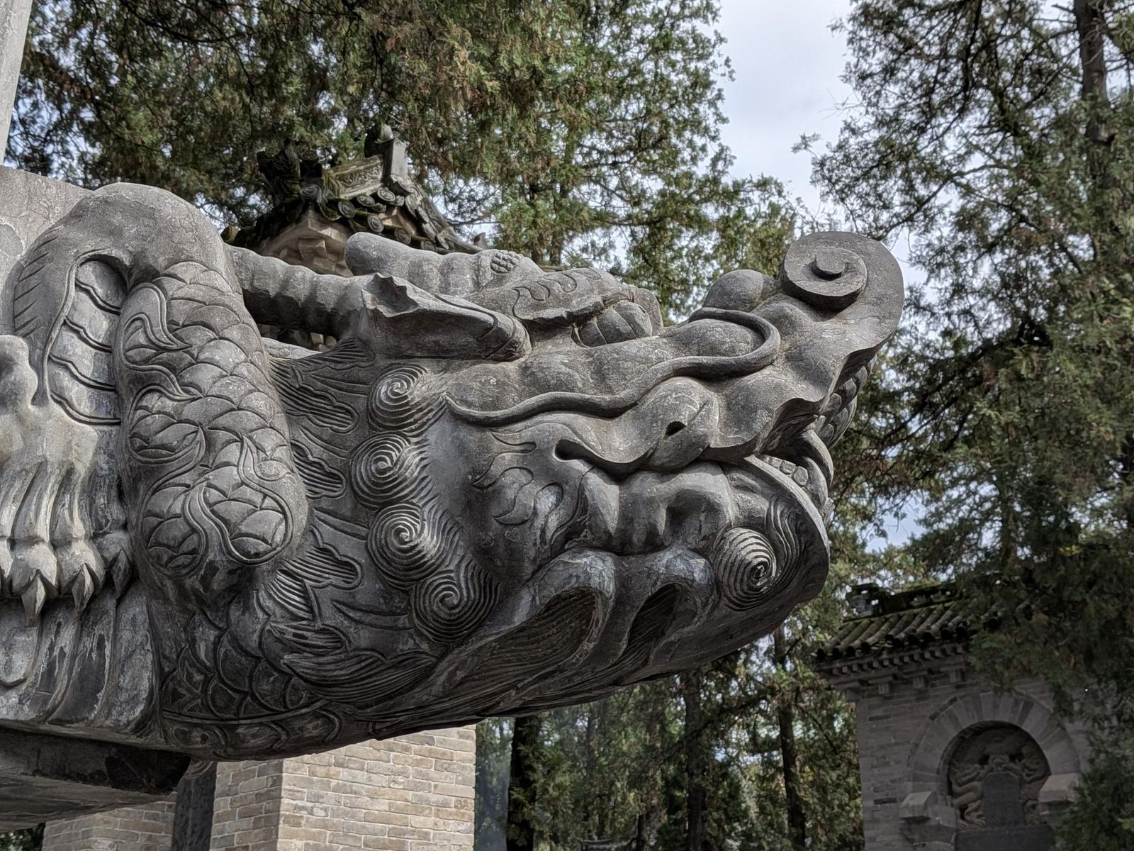 Gros plan sur une tête de dragon gravée dans la pierre, écailles finement incisées, motifs de nuages, ciel gris et cyprès en arrière-plan